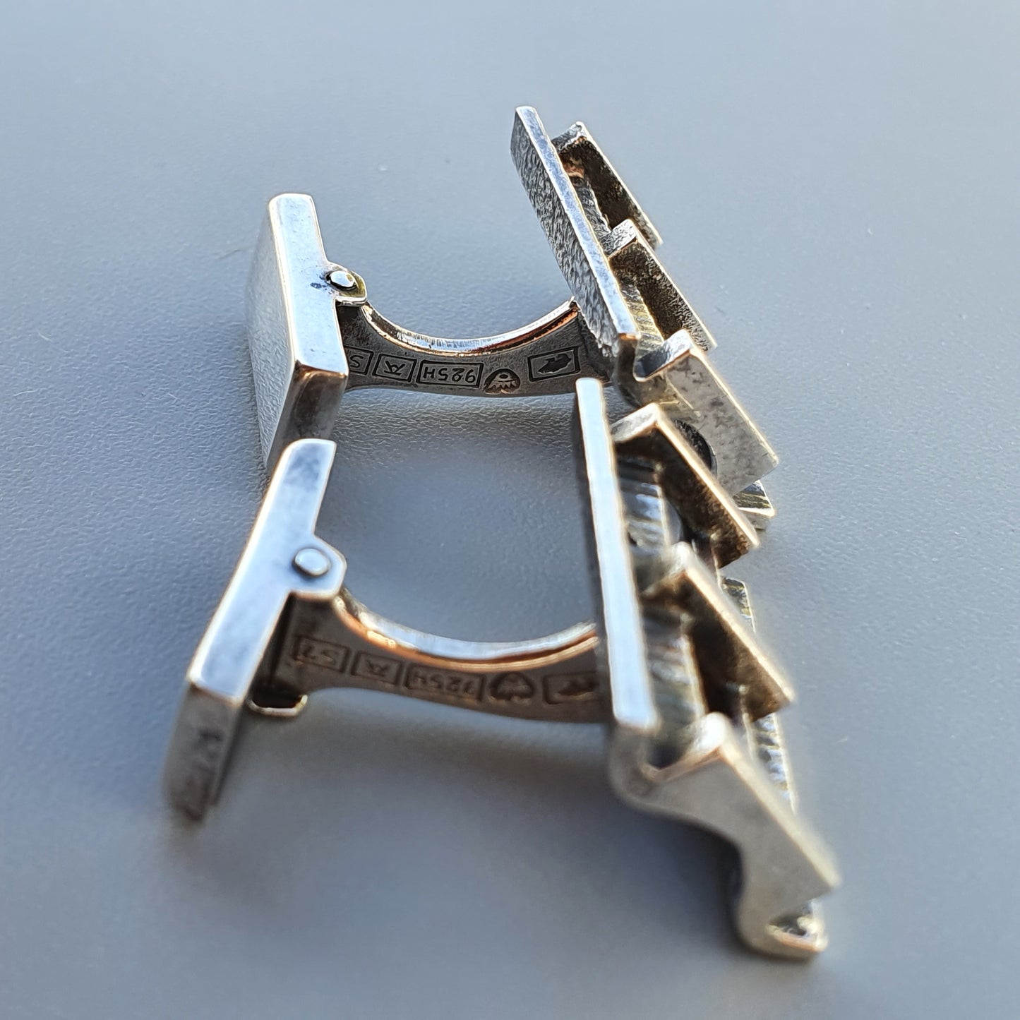 Pair of silver-colored cufflinks with decorative patterns on the bars.