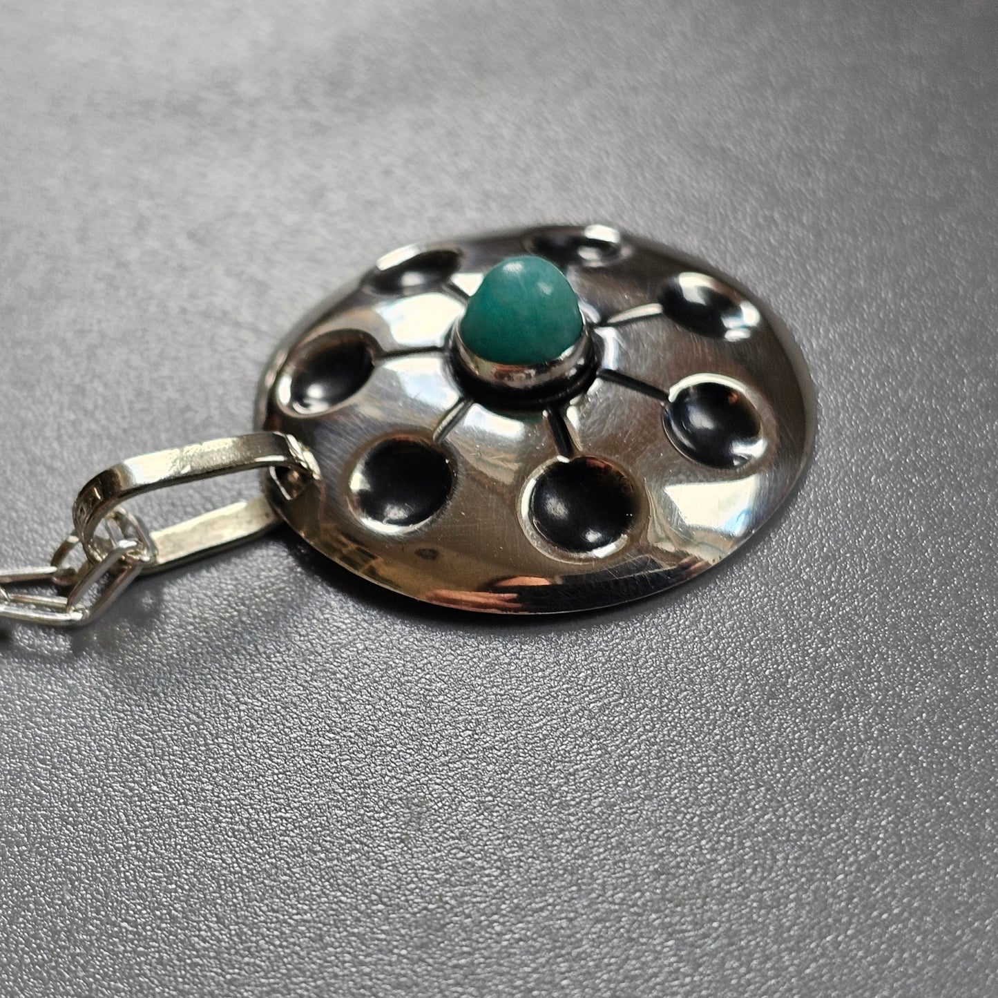 Silver pendant with a circular design featuring a central turquoise stone and surrounding holes.