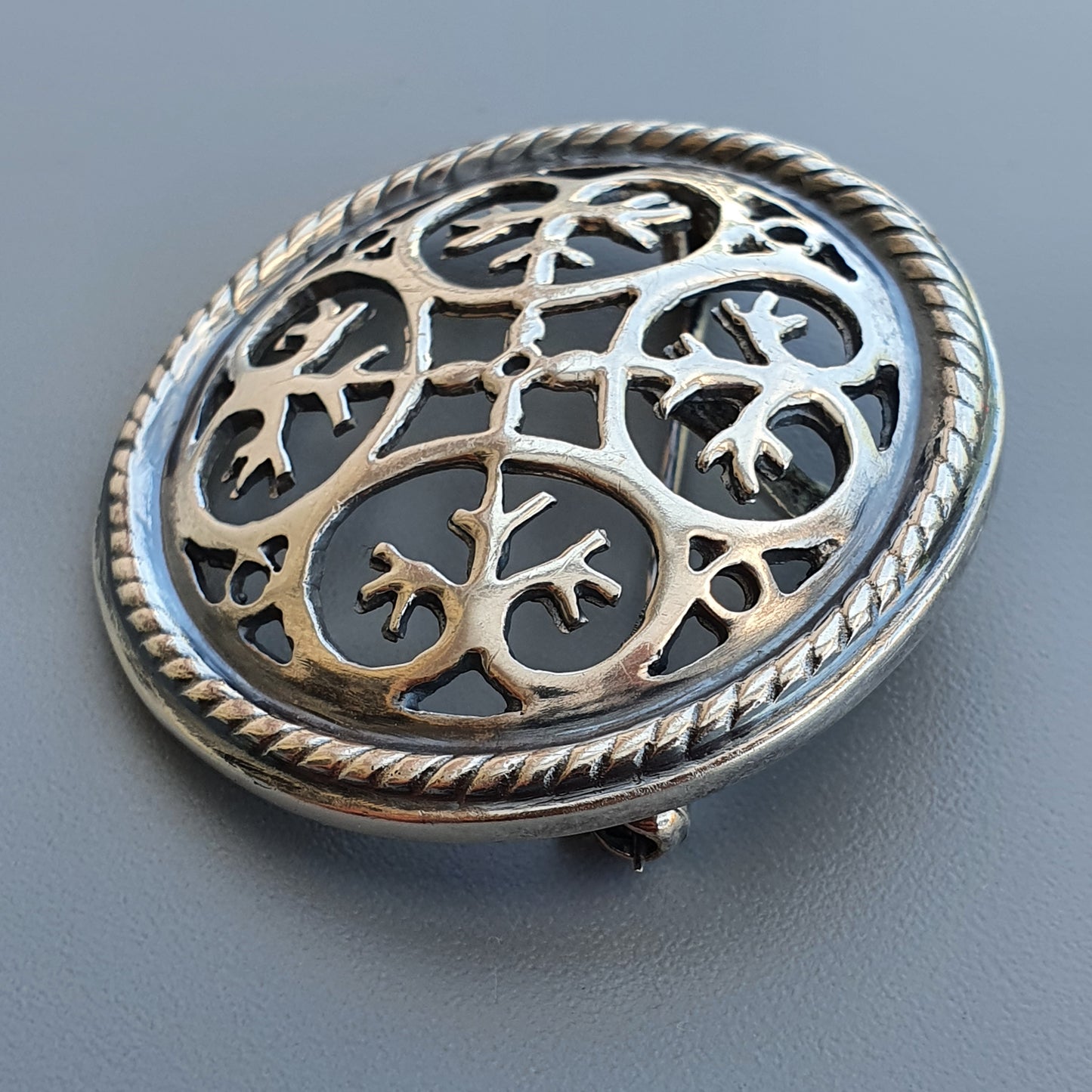 Ornate silver brooch with intricate floral or leaf-like cutout design and rope-textured border.