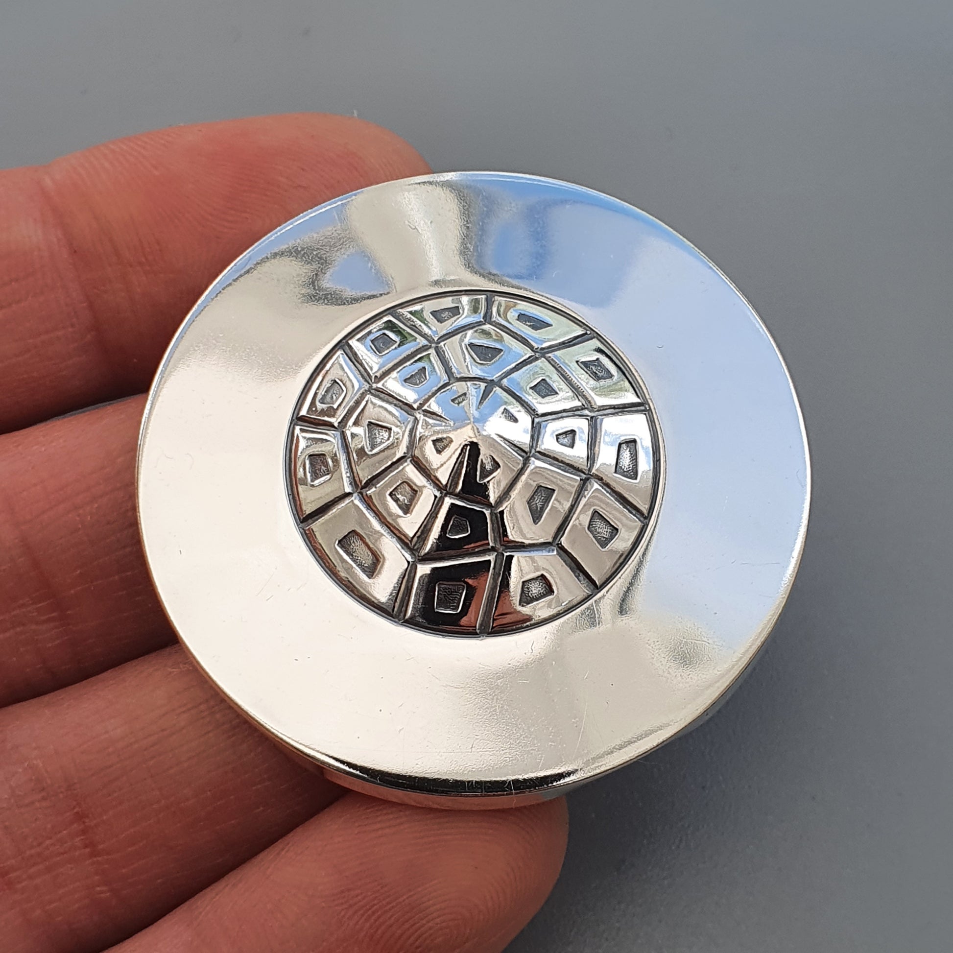 Circular silver button or pin with an intricate geometric pattern on its face.