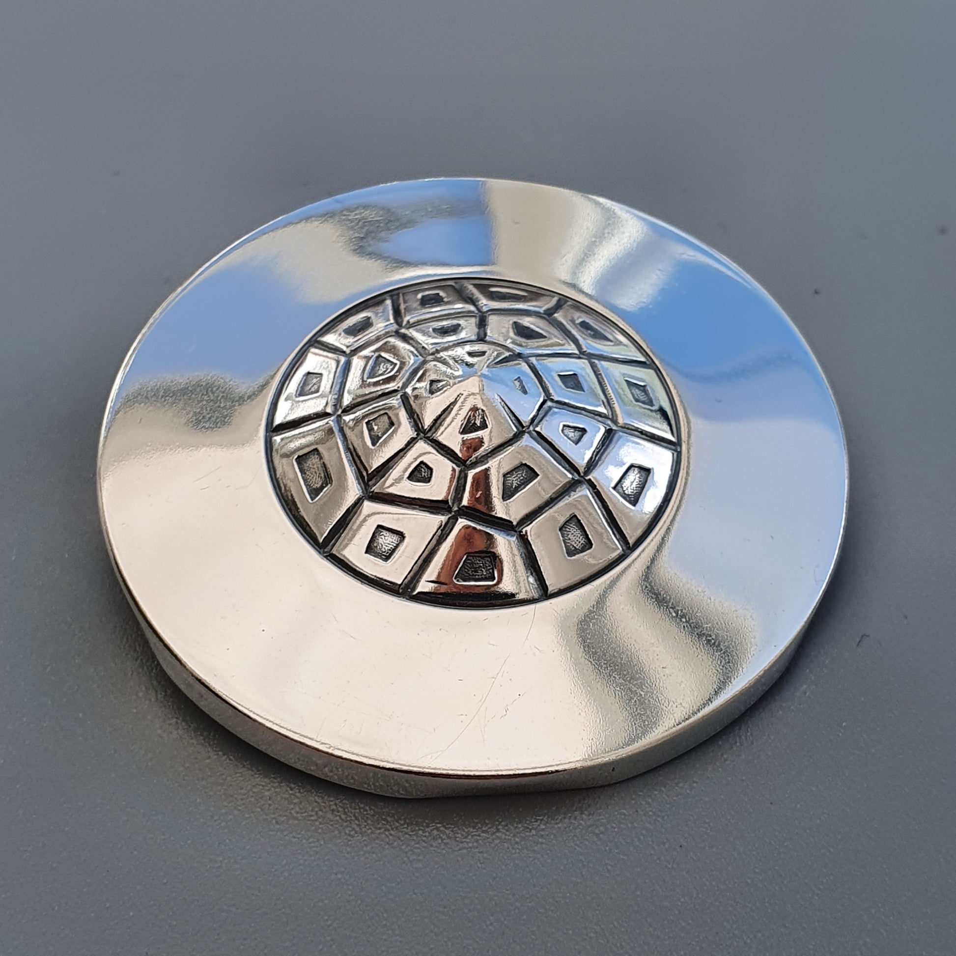 Metallic circular object with a domed center featuring a geometric pattern of shapes.