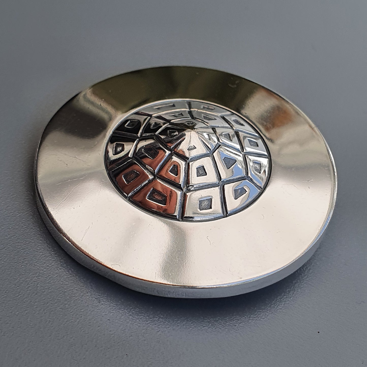 Metallic circular object with a domed center featuring a geometric pattern of small squares.