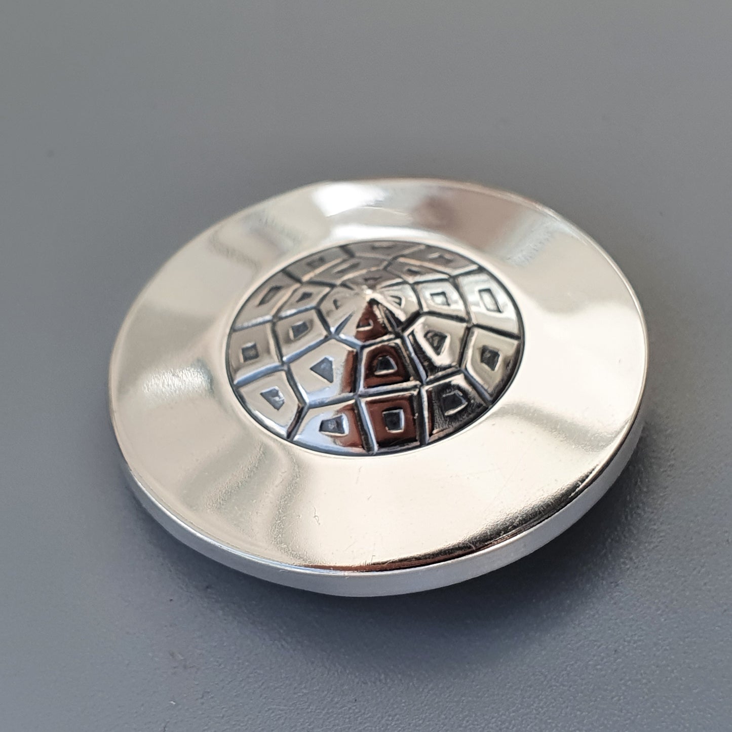 Circular silver button or fastener with a geometric pattern inlaid in the center.