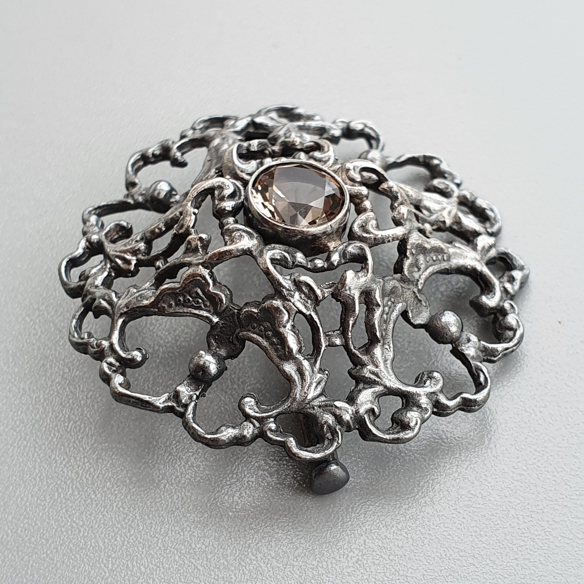 Ornate silver brooch with intricate scrollwork design and a central gemstone.