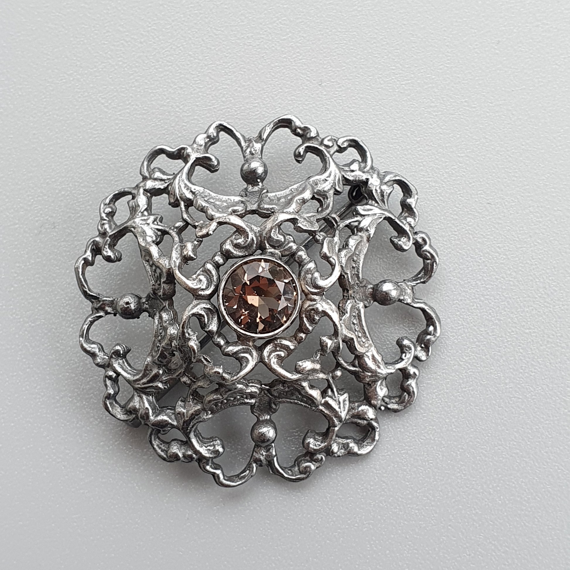 Ornate silver brooch with intricate filigree design and a central amber-colored gemstone.