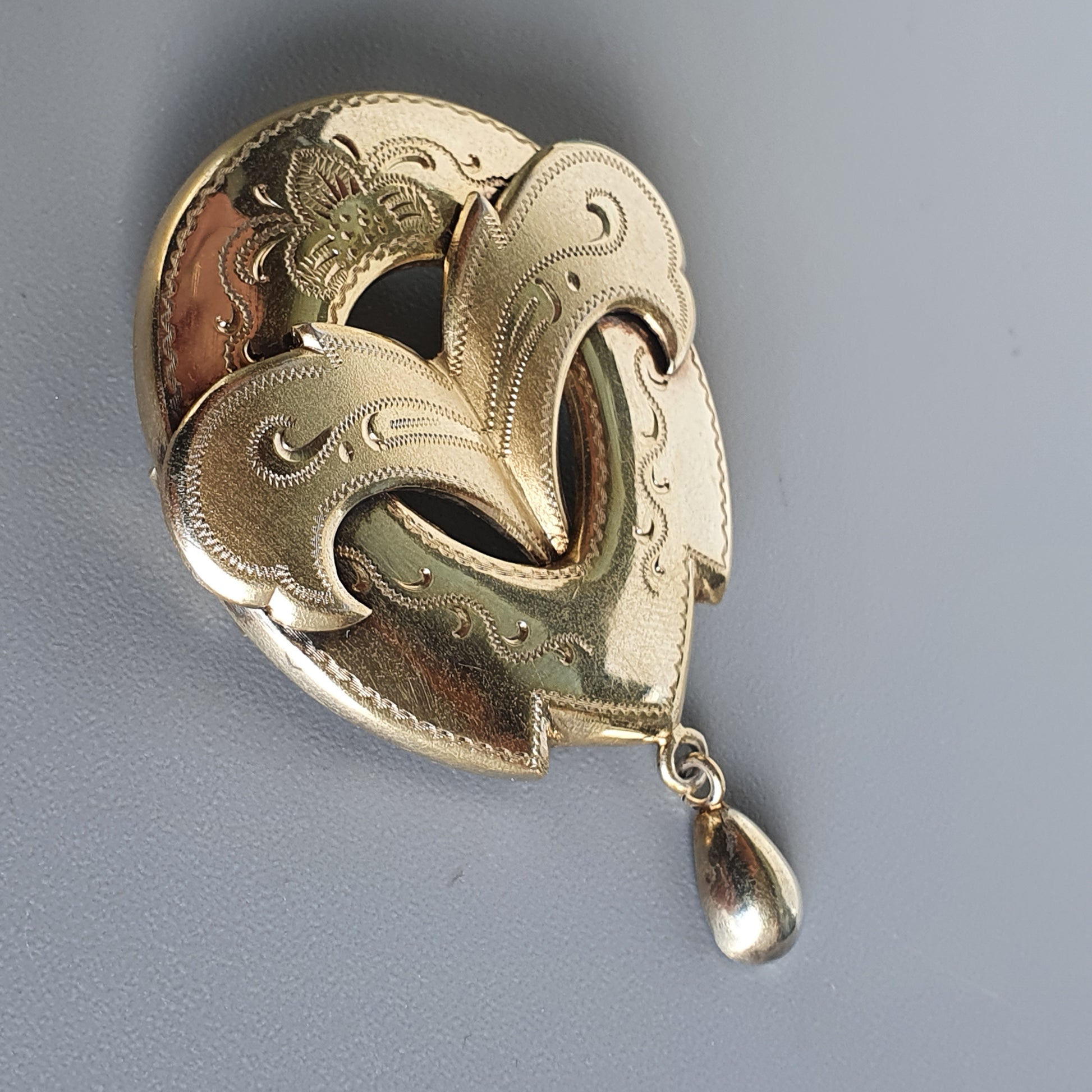 Gold heart-shaped brooch with intricate engraving and a dangling teardrop pendant.