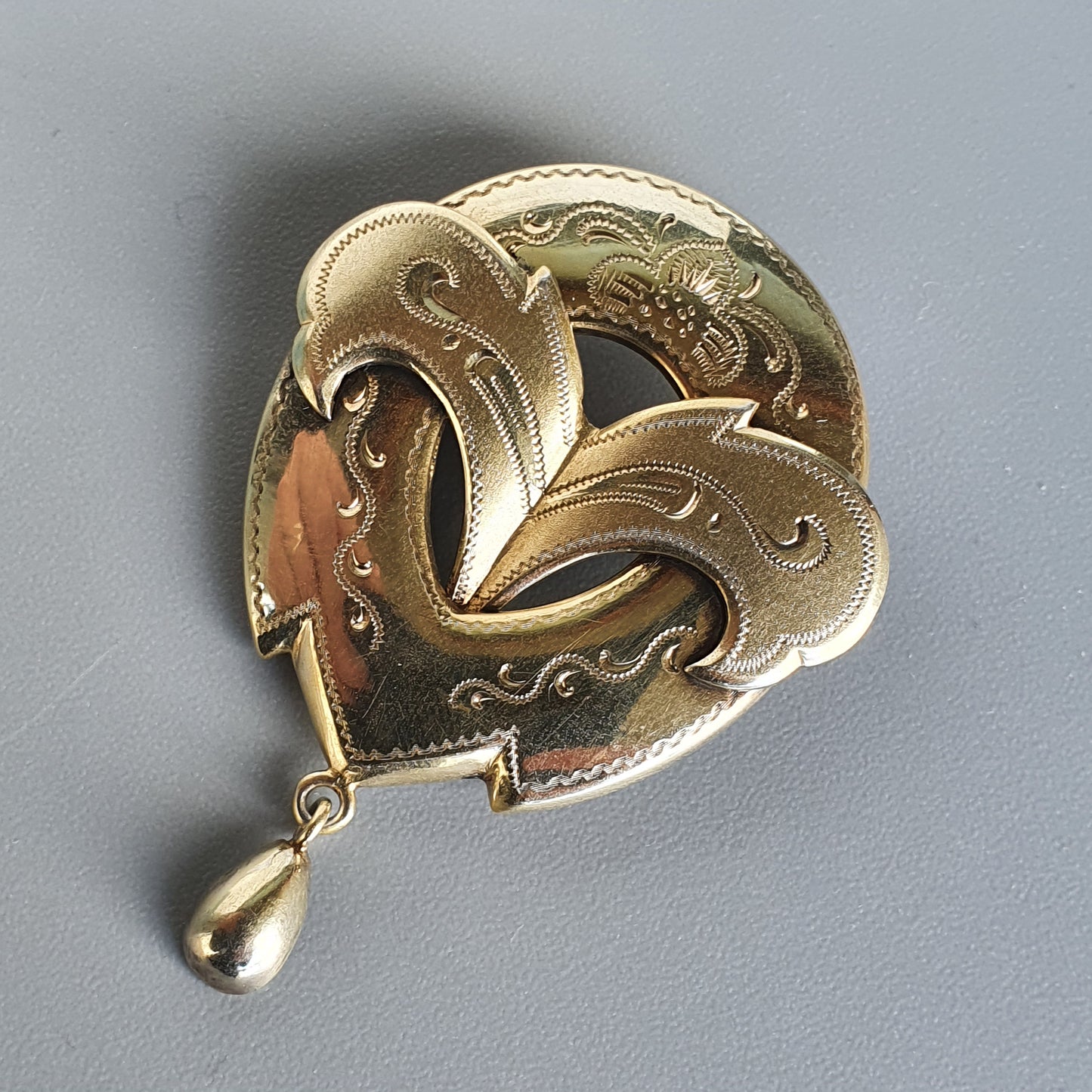 Ornate gold-toned heart-shaped brooch with floral engraving and a dangling teardrop pendant.