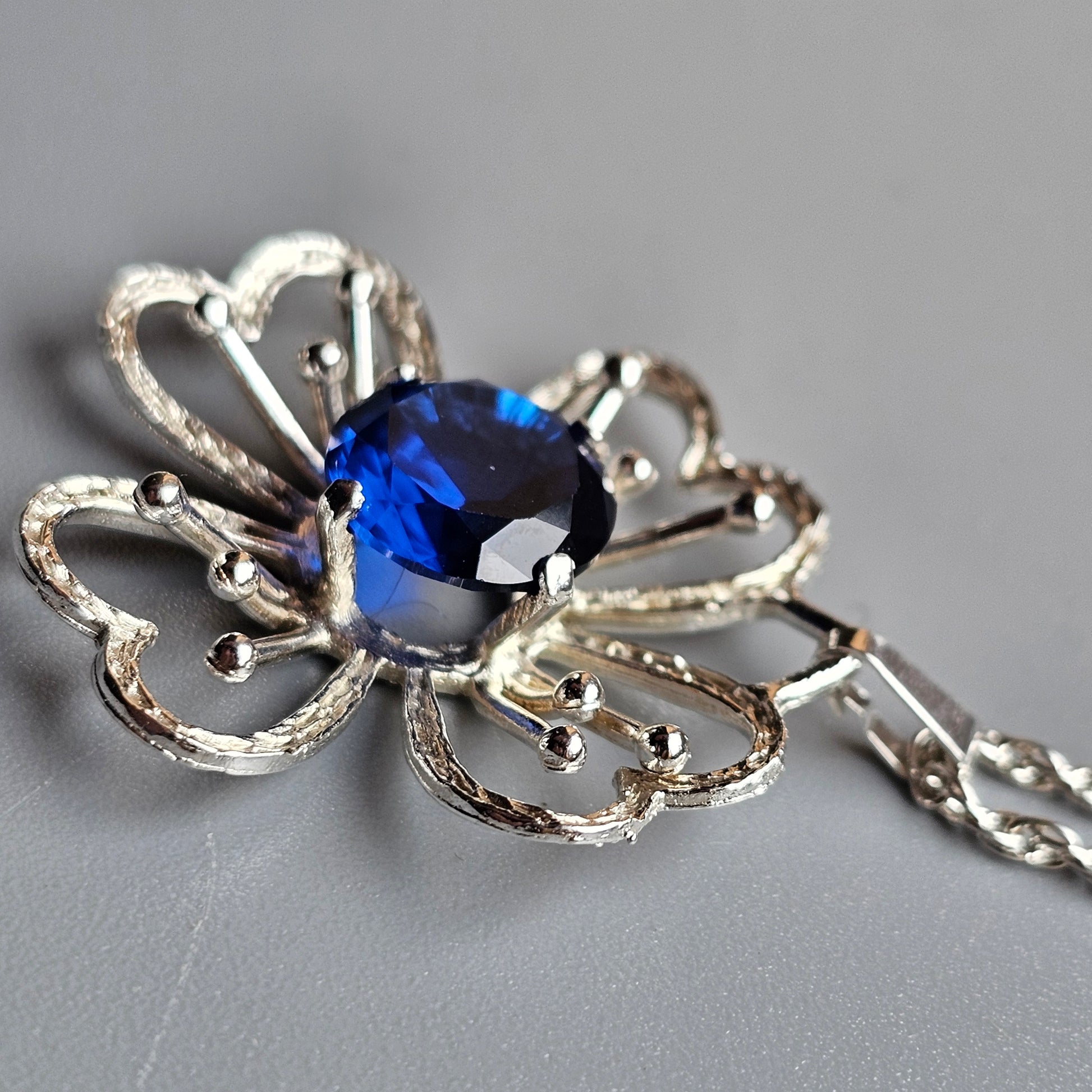 Silver pendant featuring an ornate floral design with a deep blue gemstone at its center.