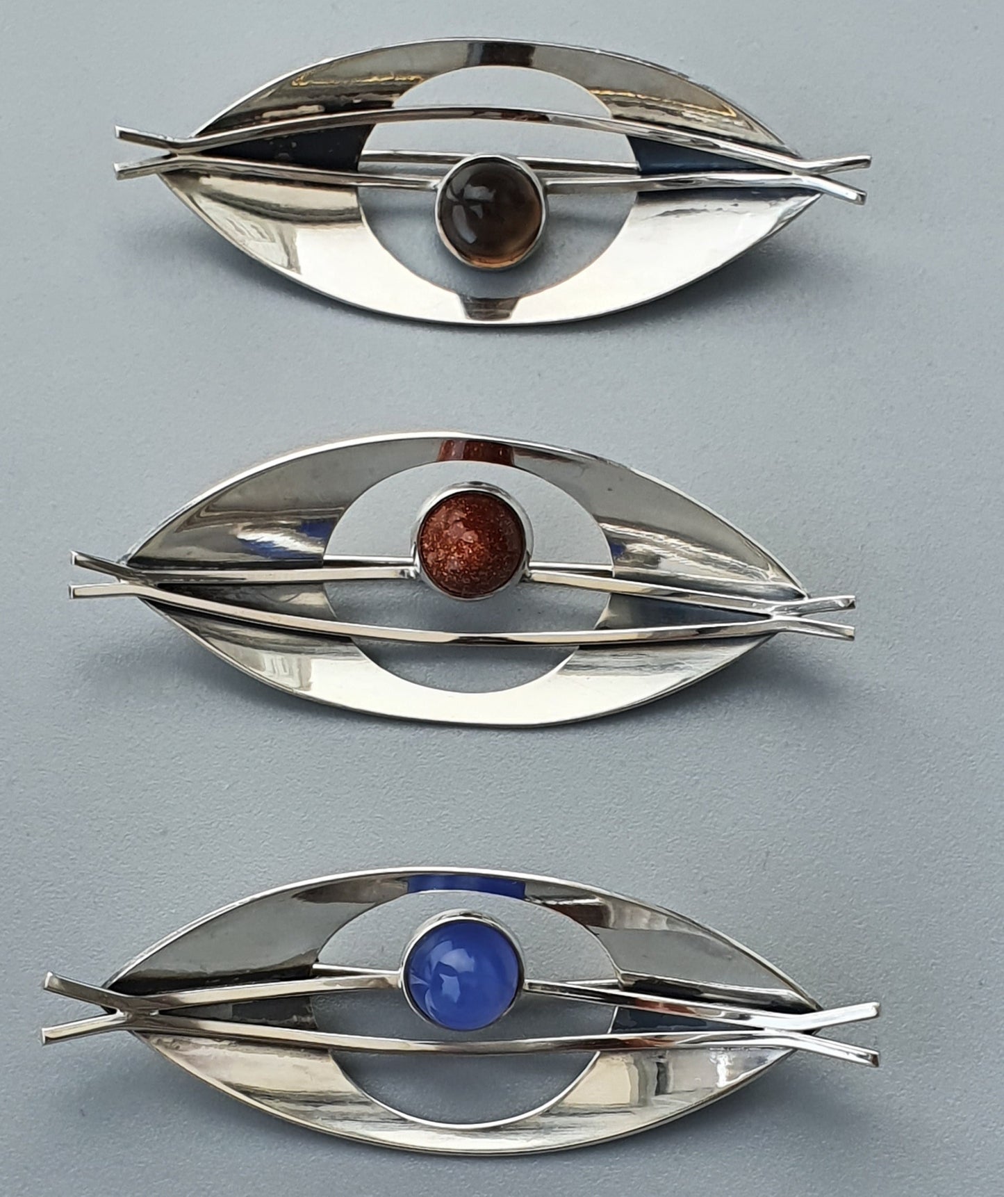 Eye-shaped silver brooches with different colored gemstones in the center.