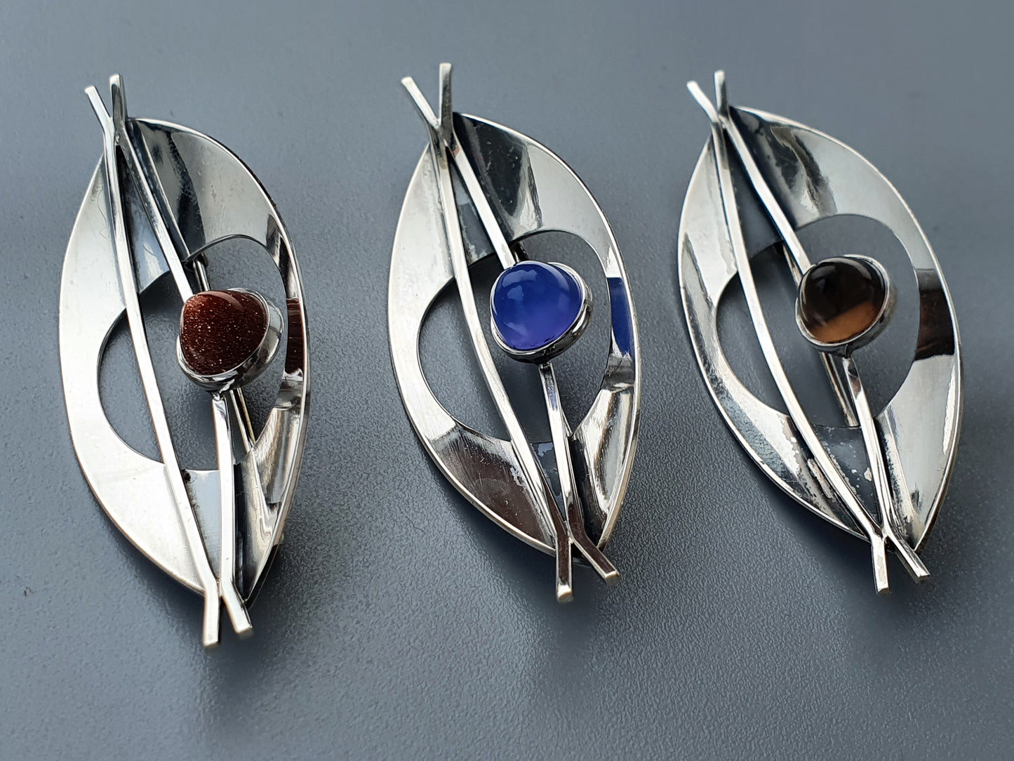 Three silver leaf-shaped brooches with colored gemstones.