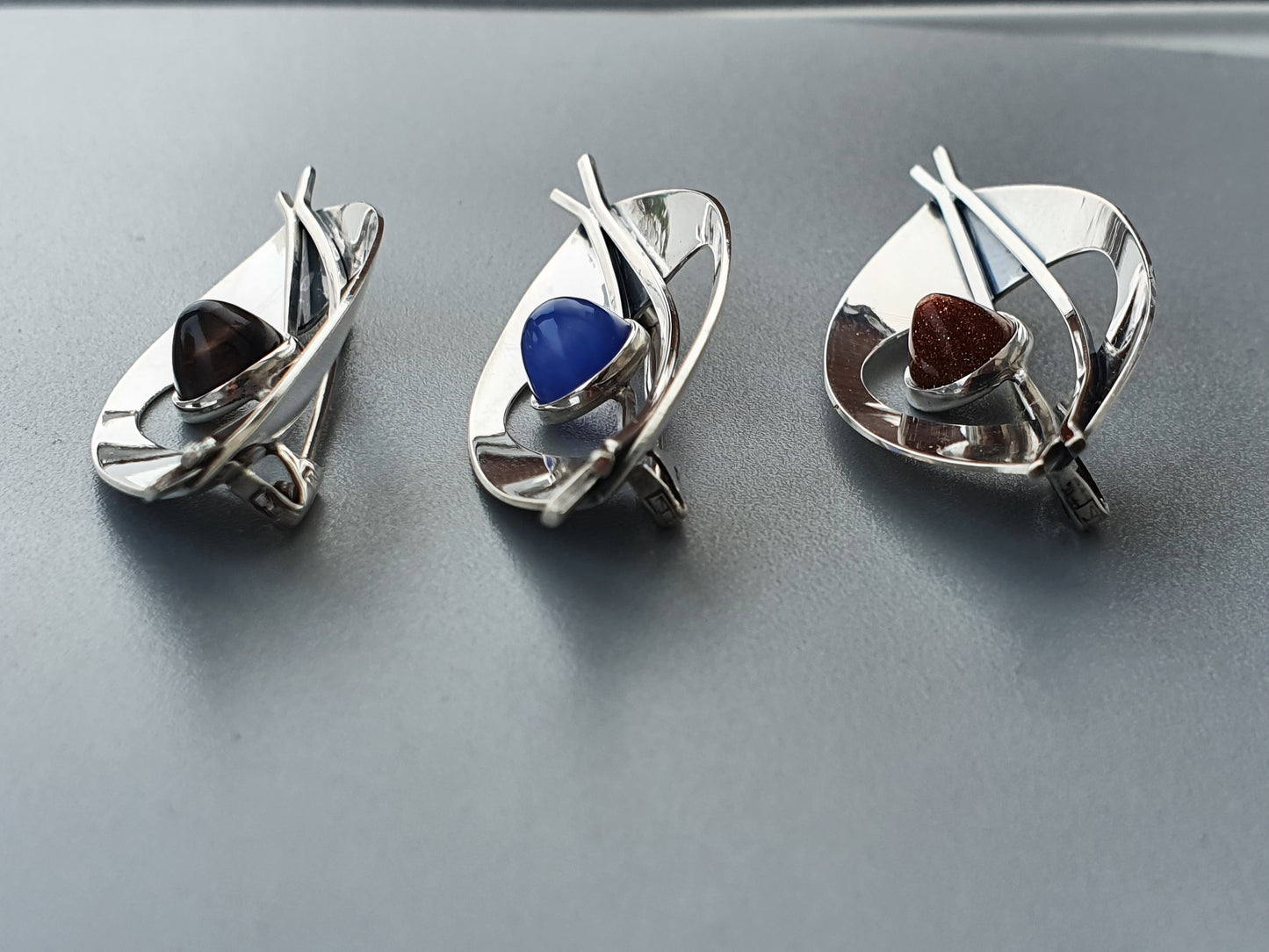 Silver-colored metal pins or brooches shaped like abstract boats, each holding a different colored stone.