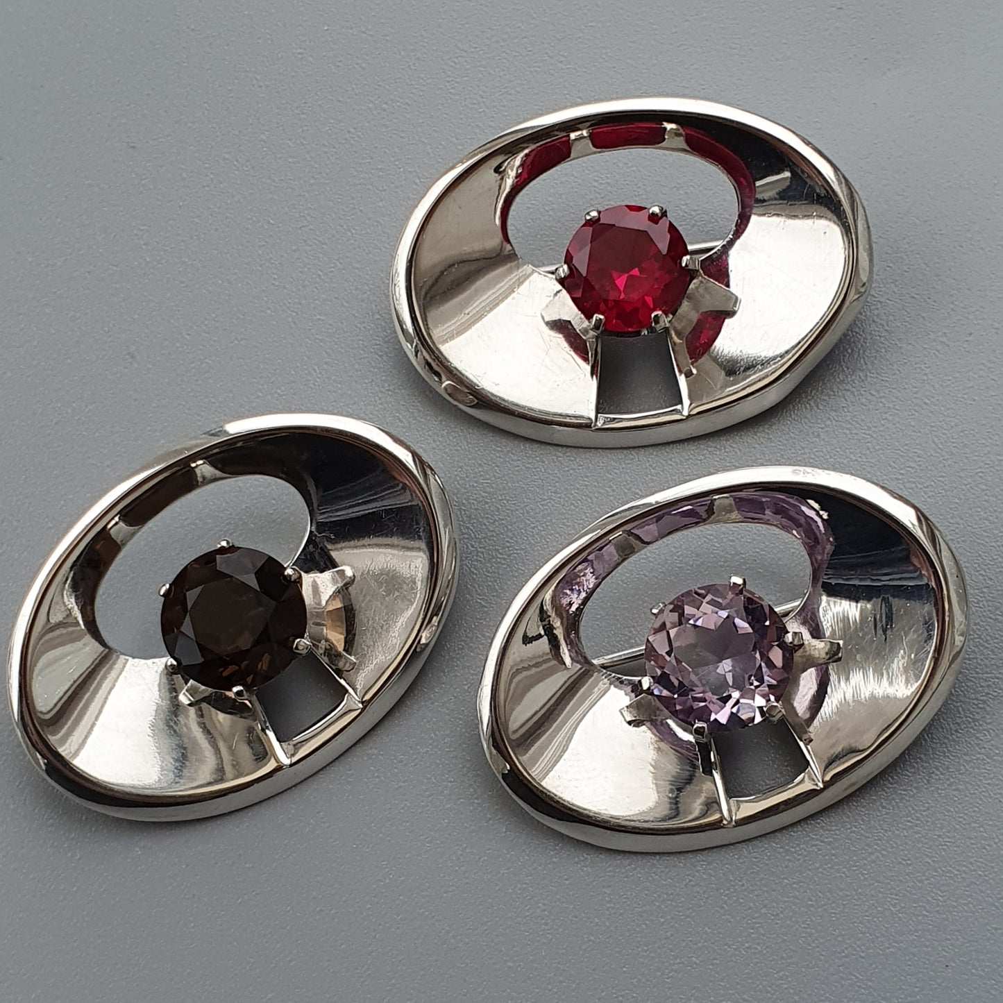 Oval silver brooches or pendants, each featuring a single colored gemstone at the center.