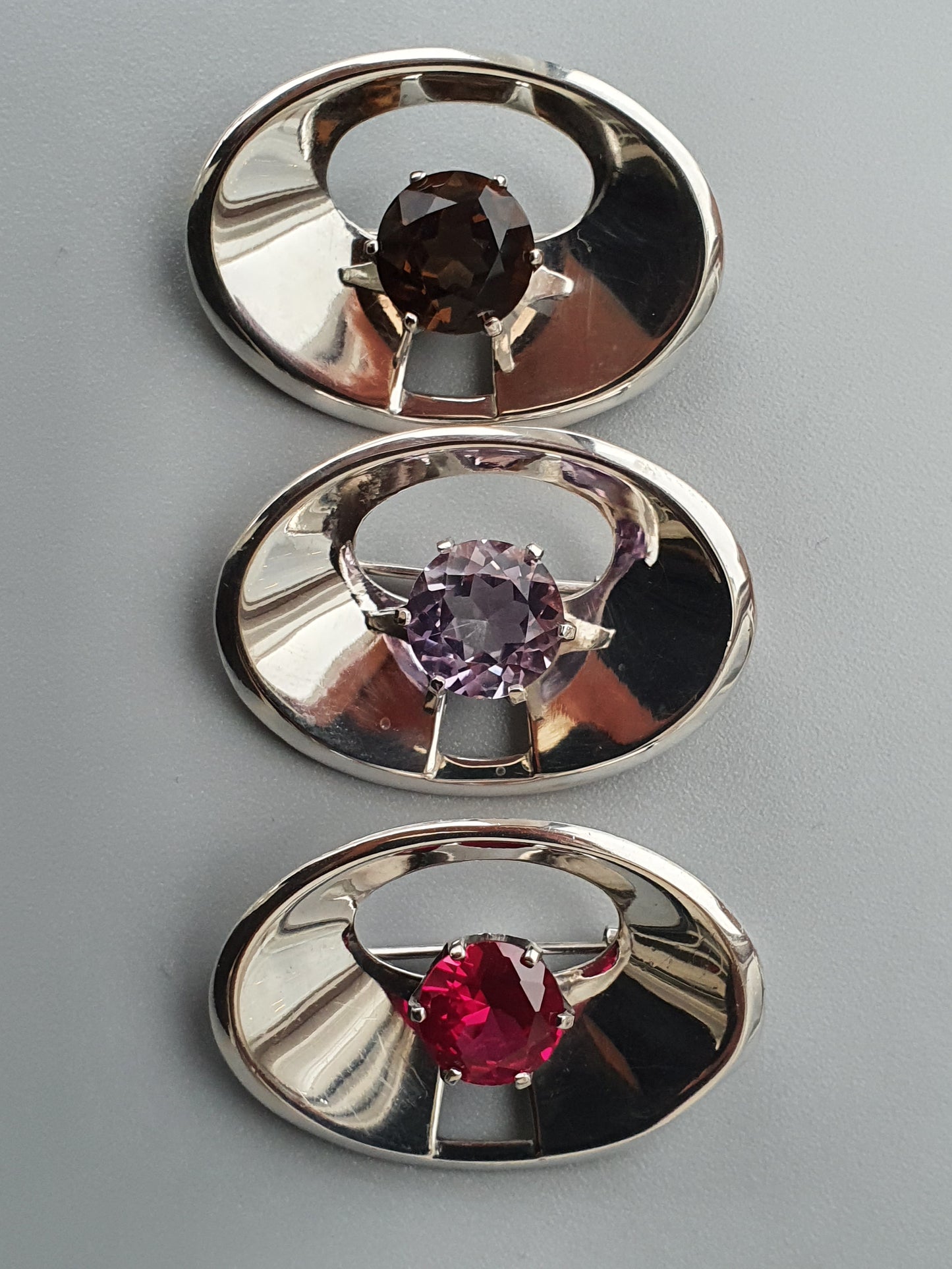 Three oval-shaped silver jewelry pieces, each featuring a different colored gemstone.