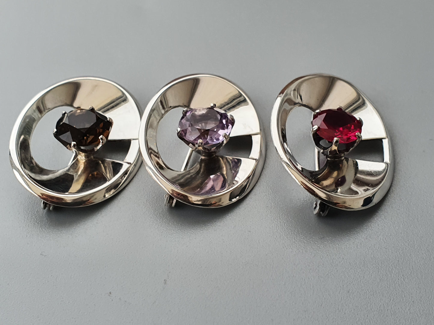 Three circular silver brooches or pins, each set with a different colored gemstone.