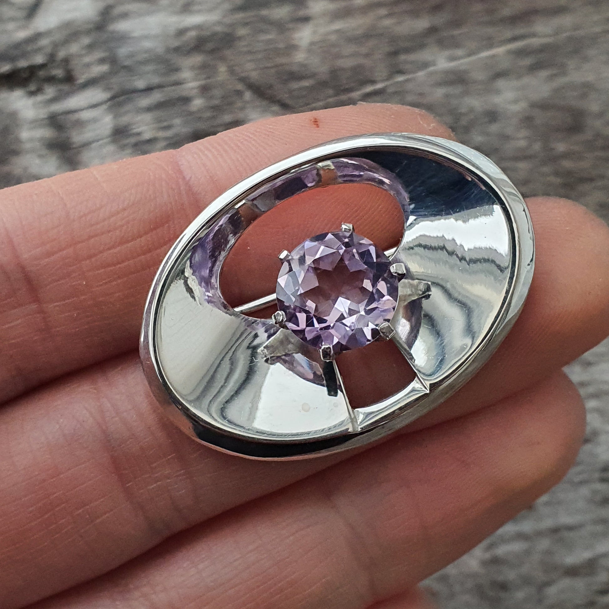 Oval silver brooch or pendant featuring a purple gemstone in a prong setting.