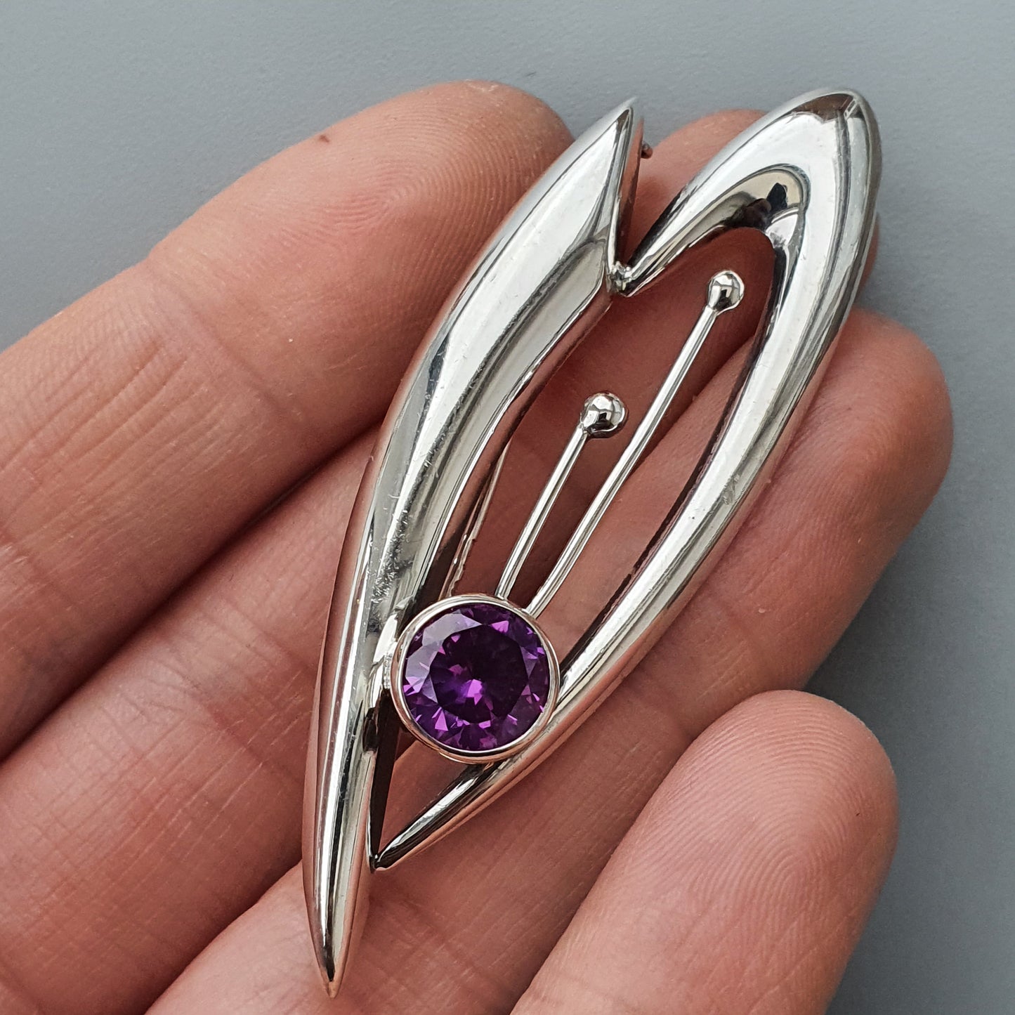 Stylized silver brooch or pendant featuring an abstract leaf-like design with a purple gemstone.