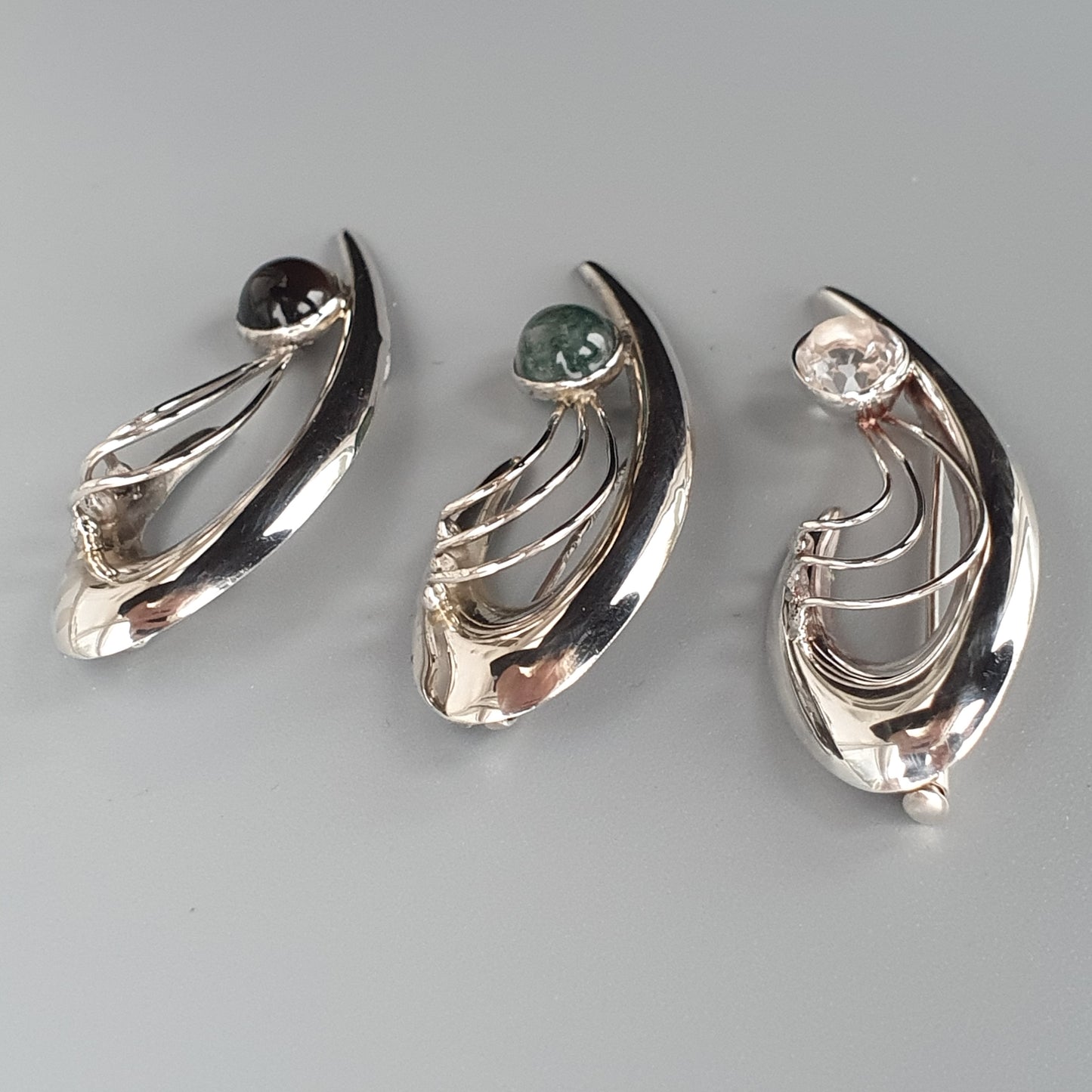 Three silver-colored abstract brooches or pendants with curved shapes and gemstone accents.