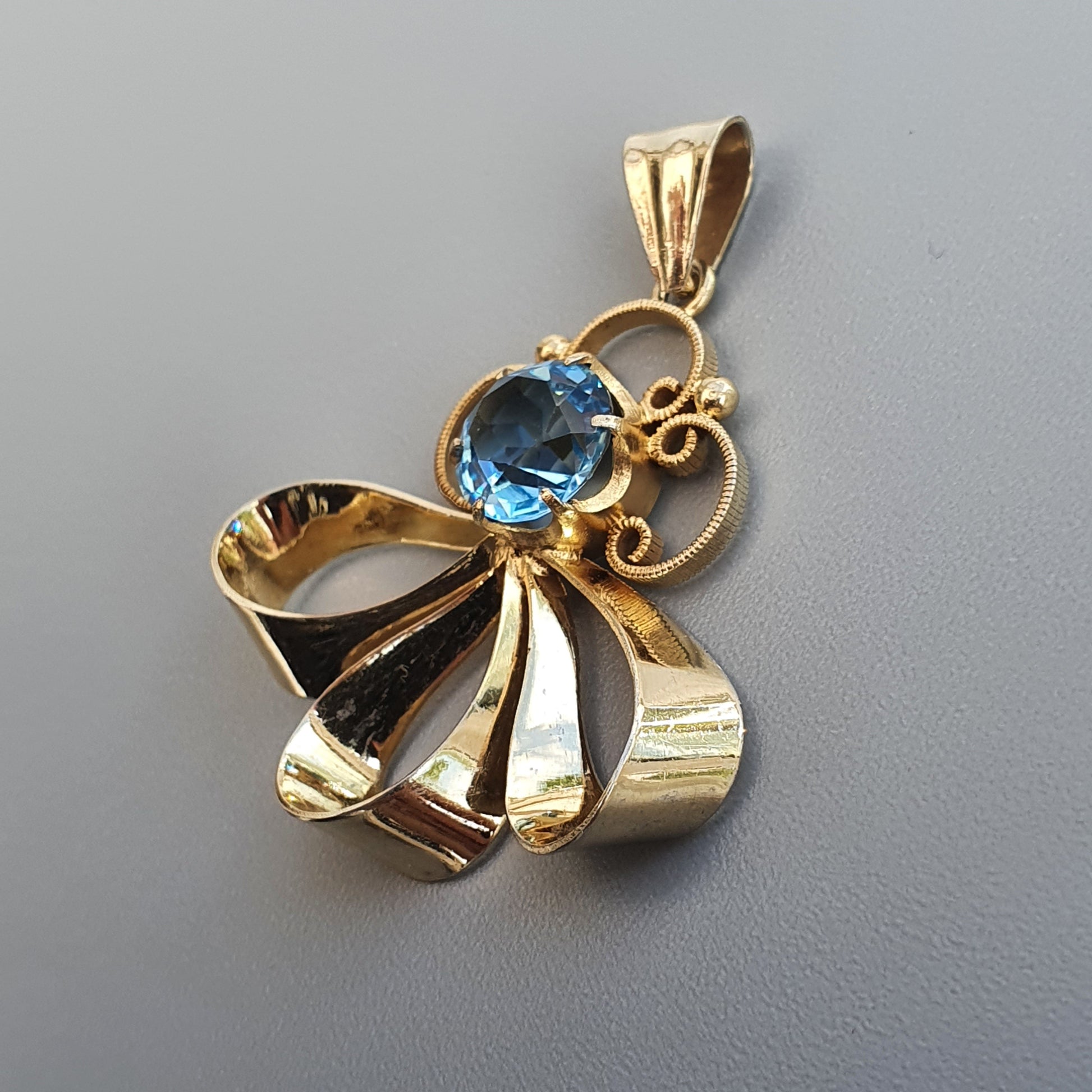 Gold pendant featuring a bow design with a blue gemstone centerpiece.