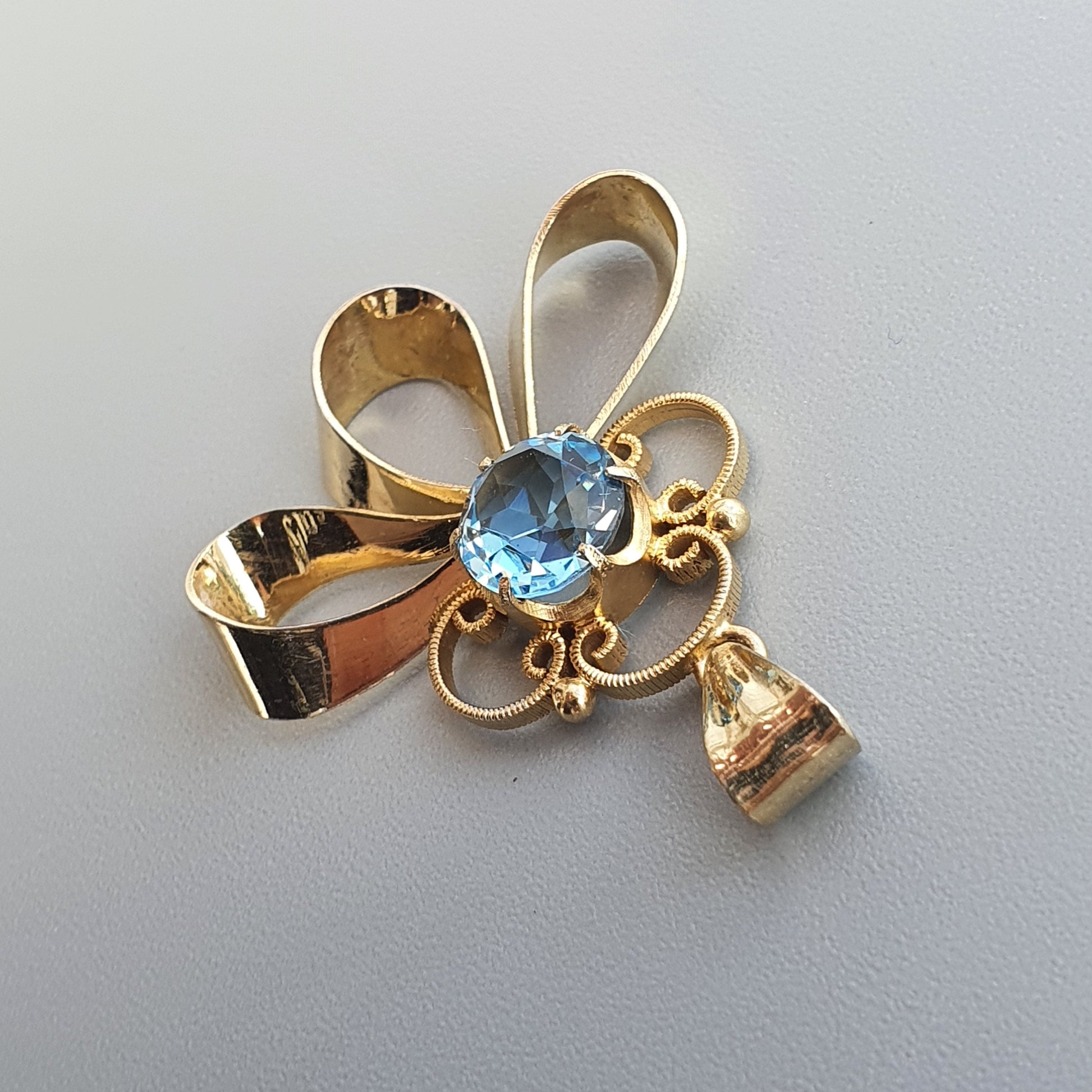 Gold-toned brooch or pendant featuring a bow design with a blue gemstone and small dangling charm.