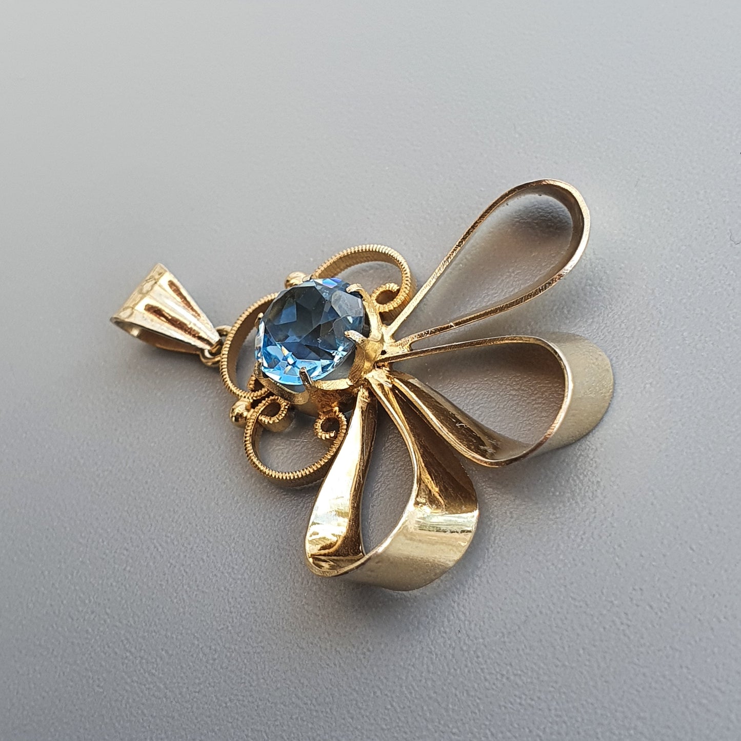 Gold pendant shaped like a bow or ribbon with a blue gemstone in the center.