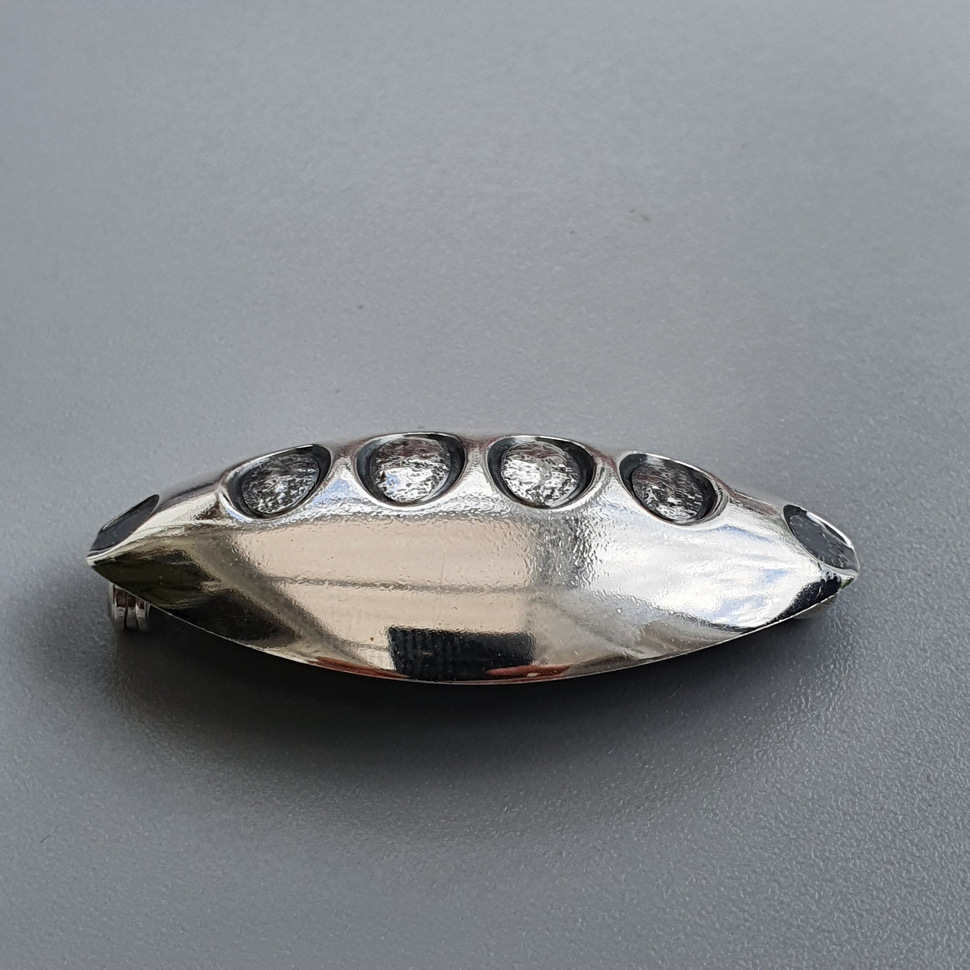 Silver-colored metallic brooch or pin with five circular indentations along its elongated oval shape.