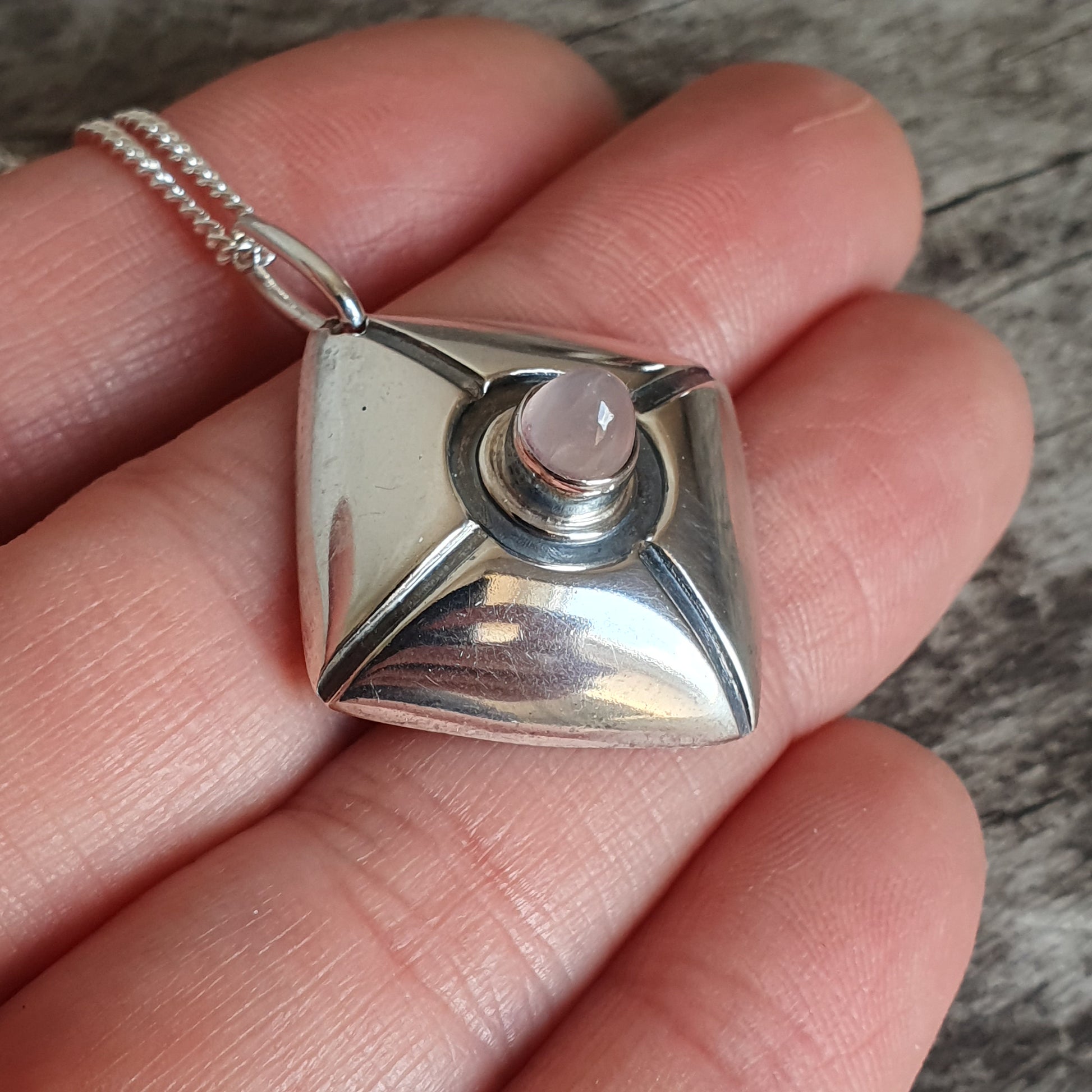 Silver pendant necklace with a square shape and a pink gemstone in the center.