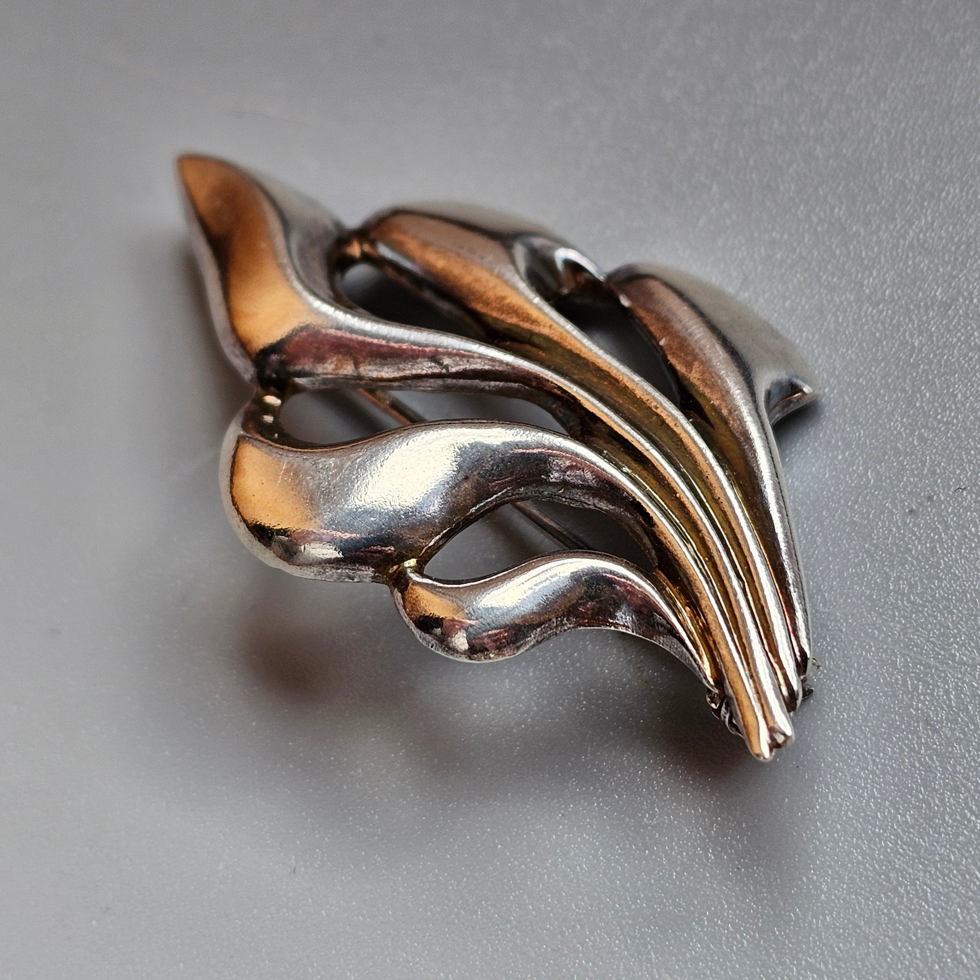 Metallic brooch or pin with a flowing, leaf-like design.