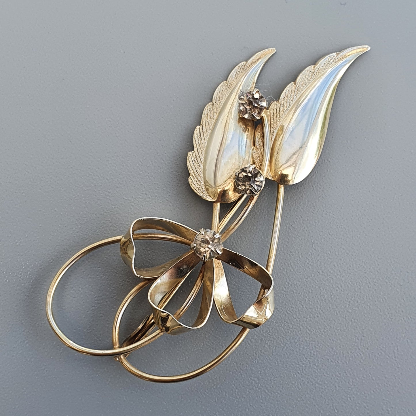 Gold-toned floral brooch featuring leaf-like shapes and curving ribbon elements adorned with small gemstones.