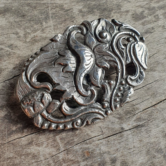 Ornate silver brooch or pendant with intricate floral and scrollwork designs.