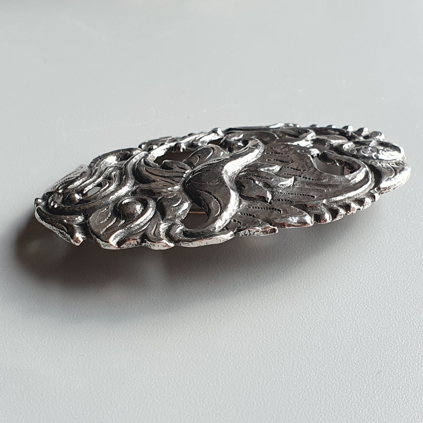 Ornate silver brooch or pin with intricate floral and swirling designs.