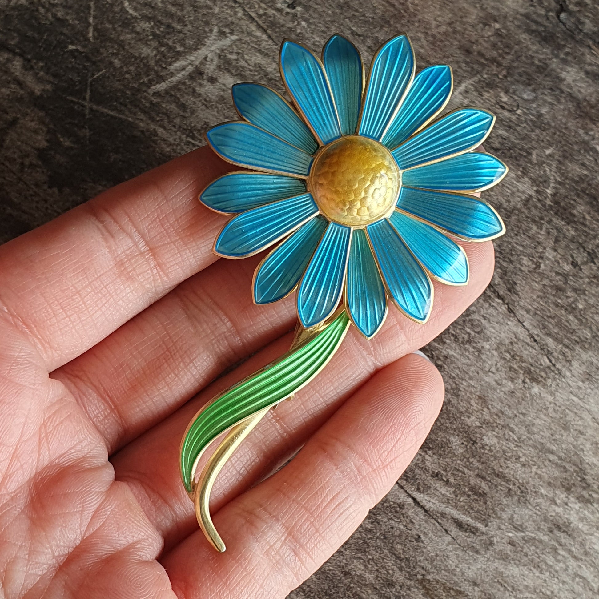 Vibrant blue and yellow enamel daisy brooch with a green stem held in a hand.