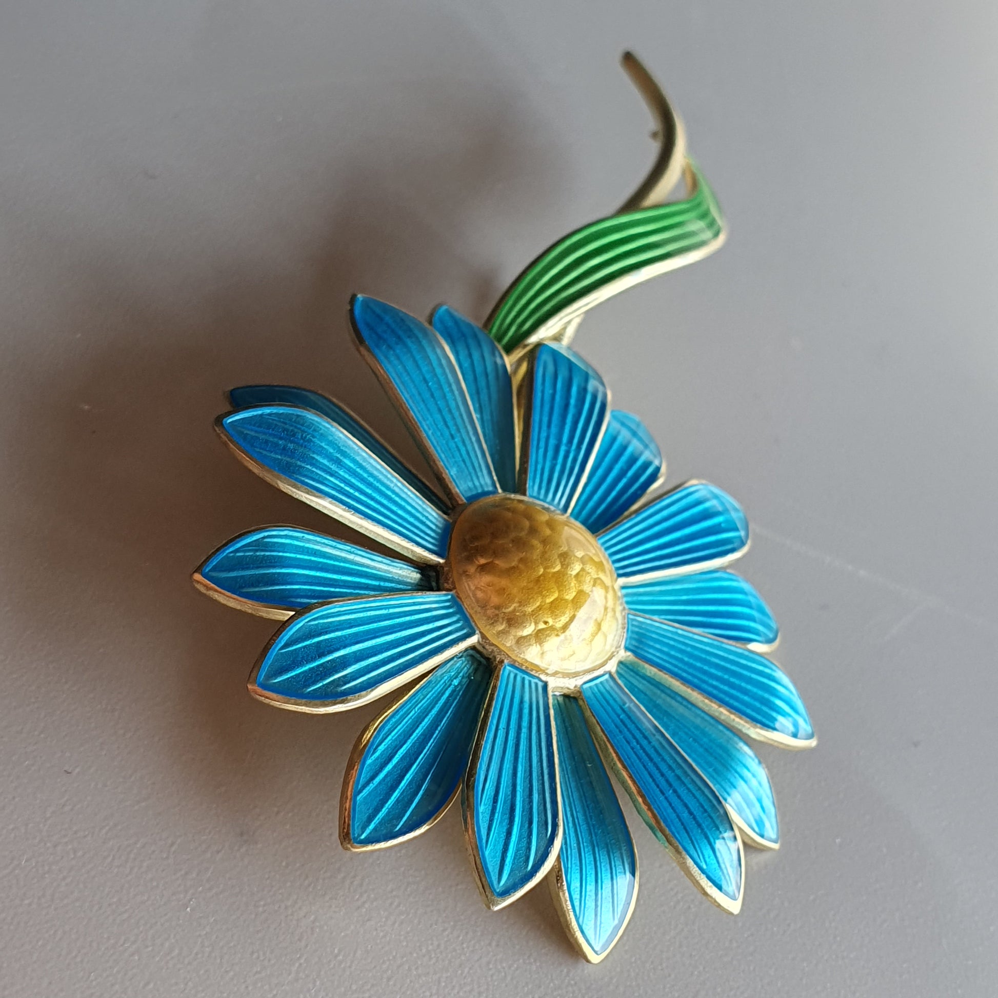 Bright blue enamel flower brooch with a yellow center and green leaf detail.