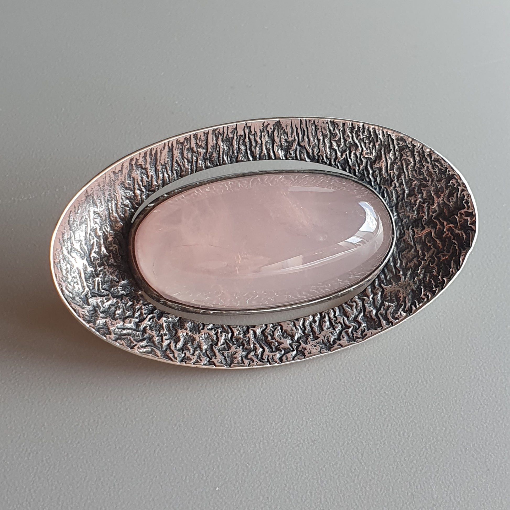 Oval brooch with a textured silver border surrounding a smooth pink gemstone or cabochon.