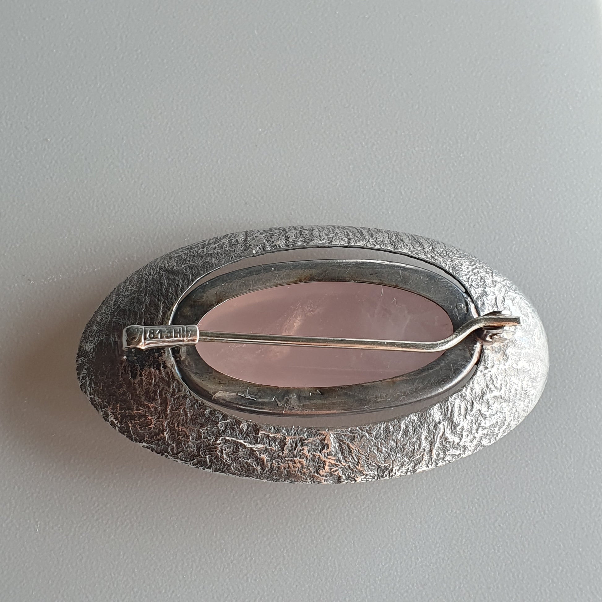 Oval silver brooch with a textured surface and pink gemstone or enamel center.