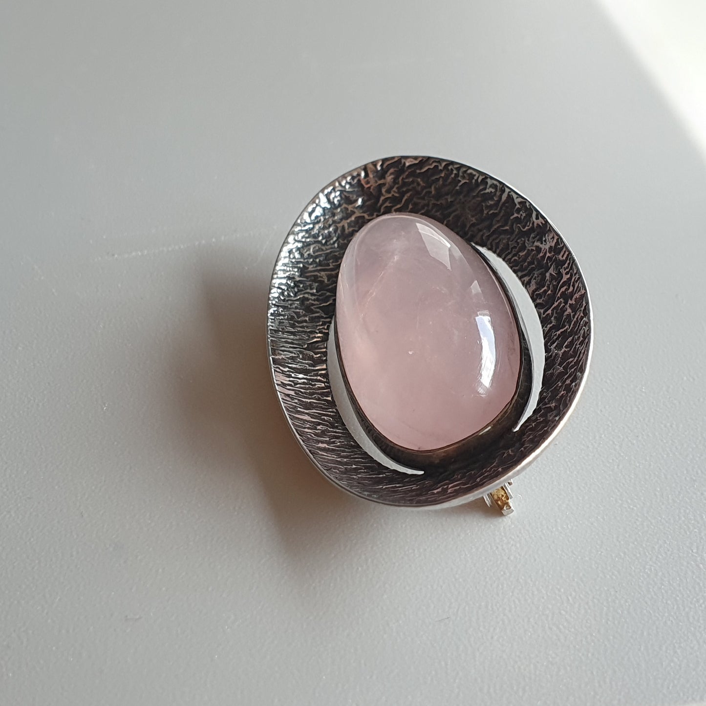 Silver brooch or pendant featuring a smooth, oval pink gemstone set in a textured circular frame.