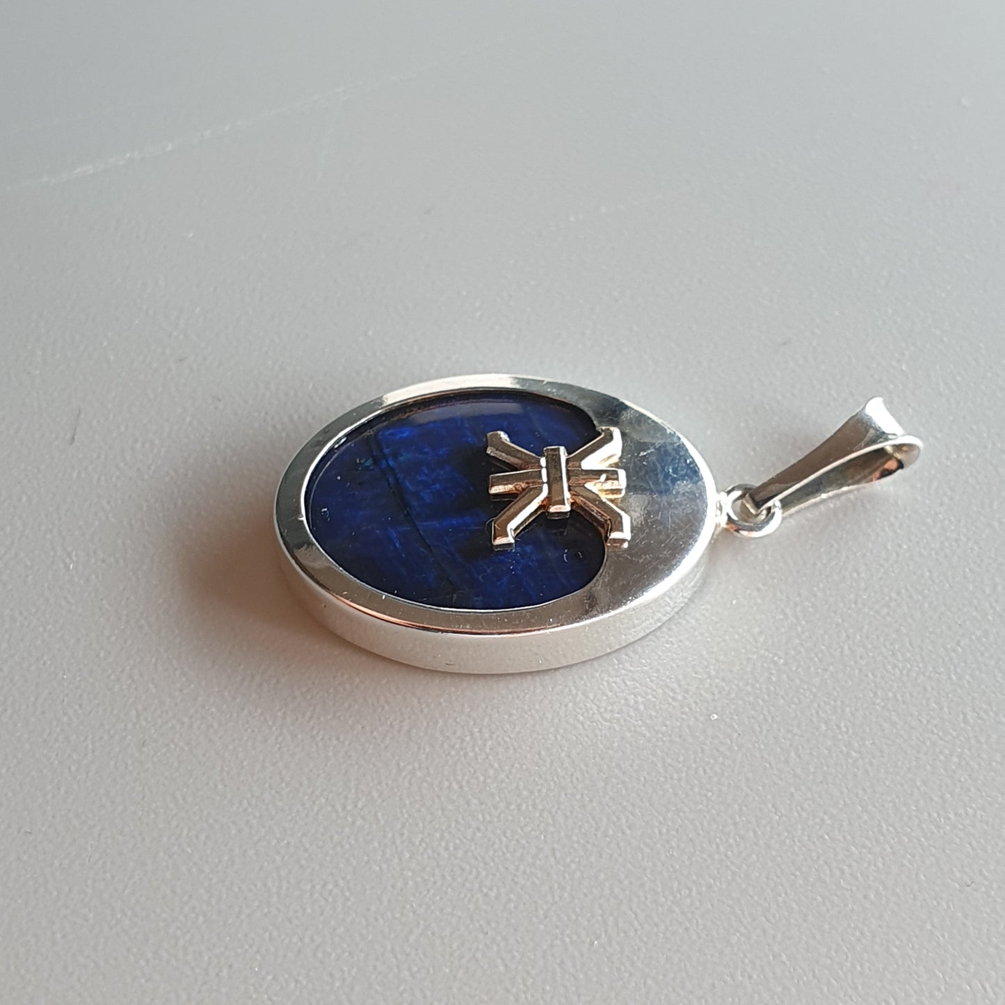 Oval silver pendant with a blue stone or enamel center featuring a small gold bow design.