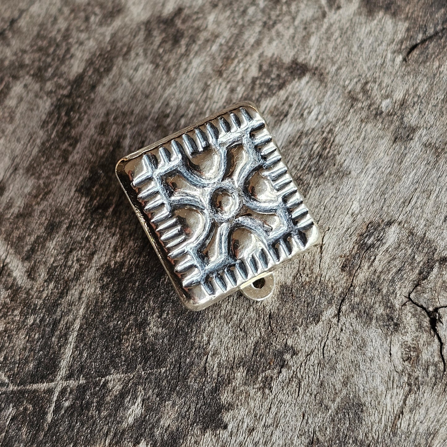 Square silver ornament with a decorative cross pattern and notched edges.