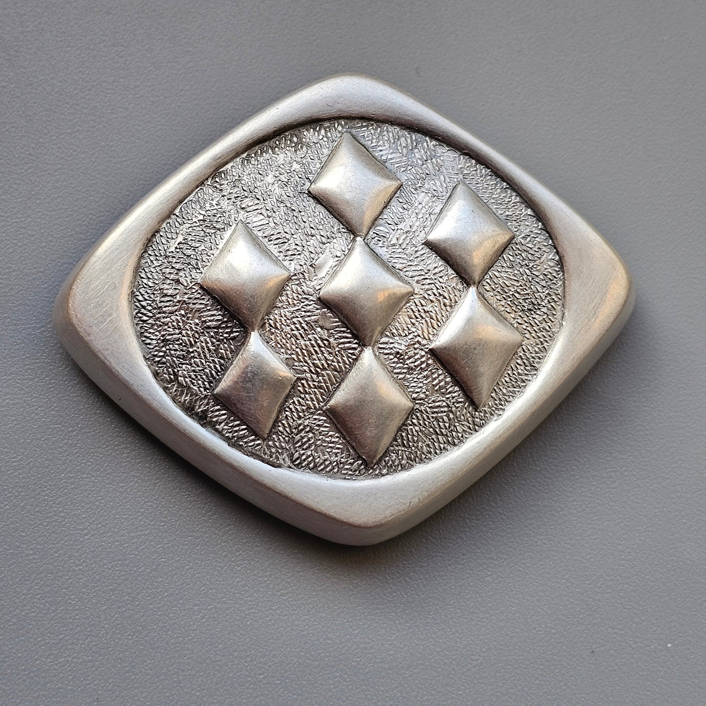 Silver brooch or pendant with a diamond-shaped pattern of raised squares on a textured background.