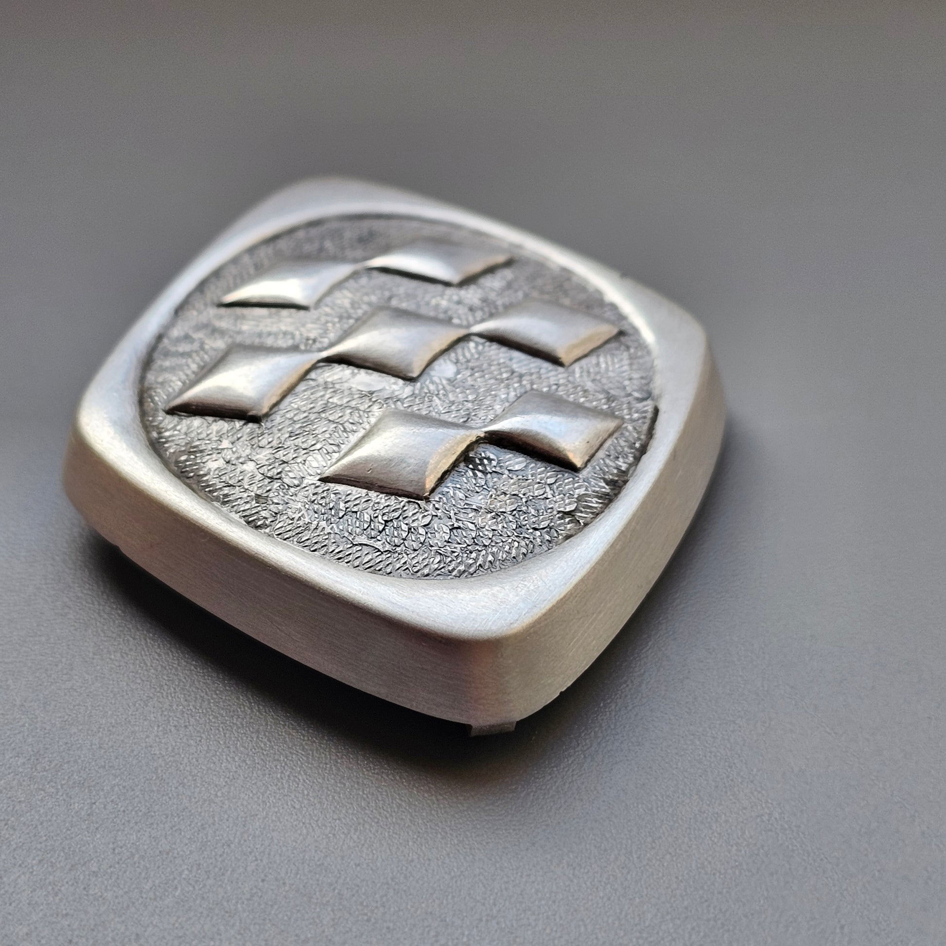 Silver-colored metal emblem with a geometric pattern of raised squares.