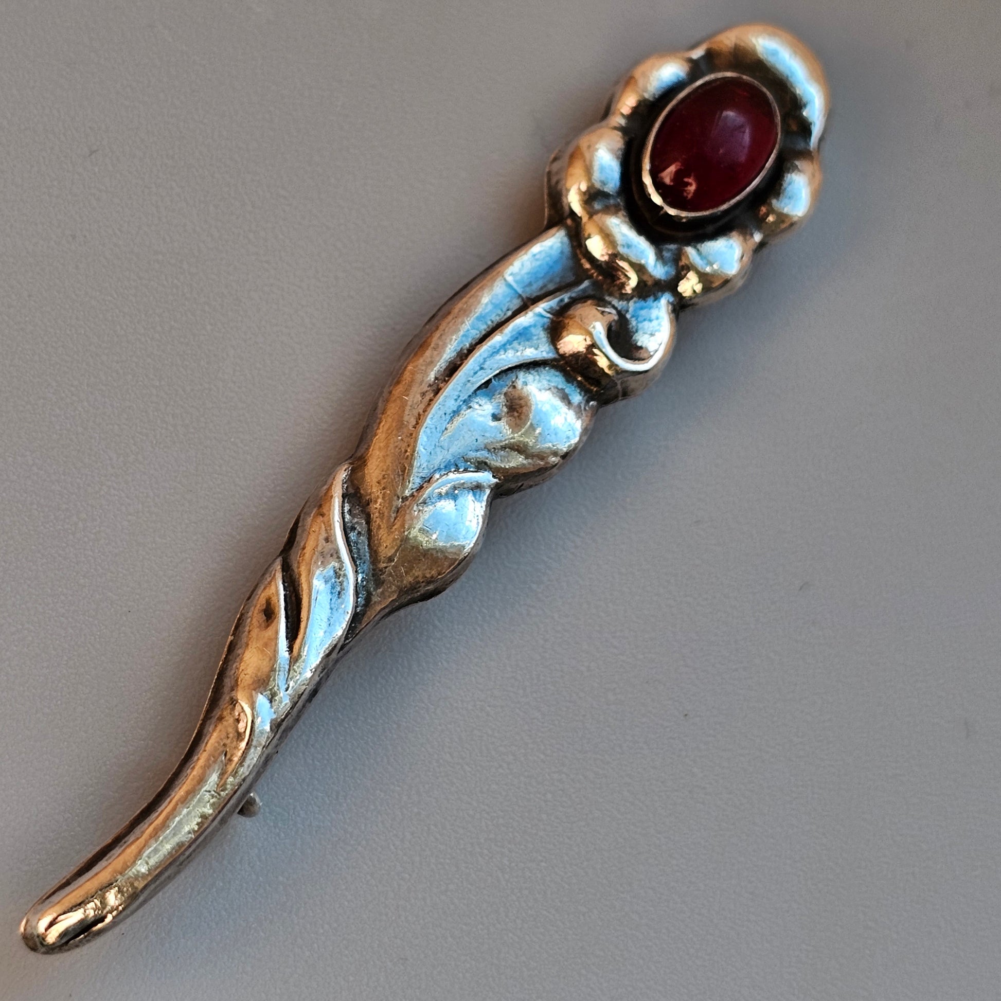 Ornate silver brooch with a curved shape and a central red gemstone.