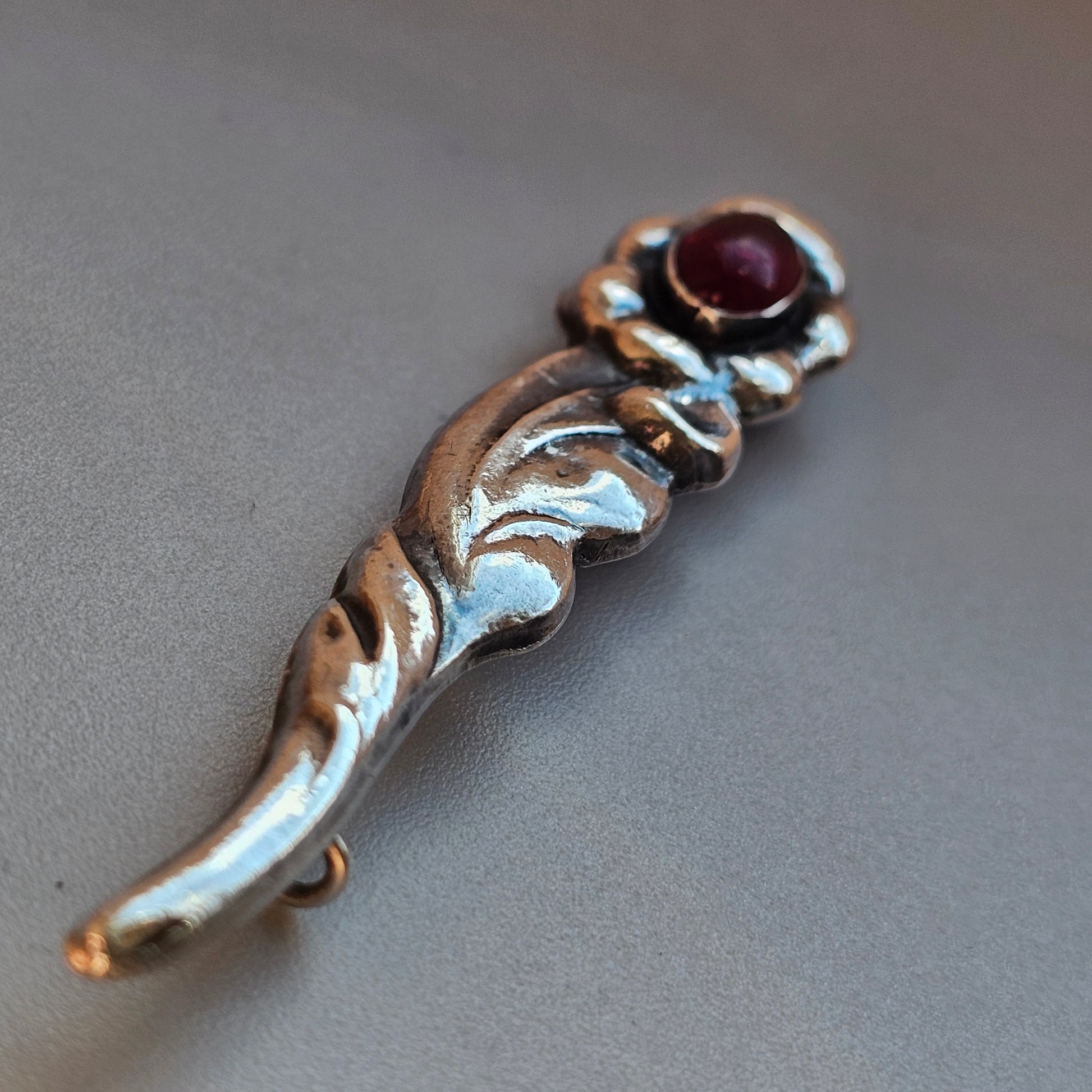 Ornate silver brooch or pin with a curved leaf-like shape and a small red gemstone.