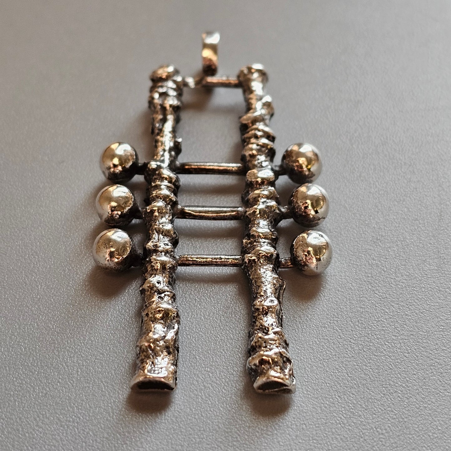 Silver or metal pendant shaped like bagpipes with decorative spheres and texturing.