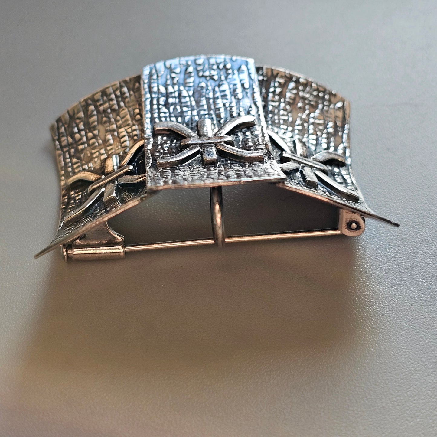 Ornate silver brooch with textured panels and bow designs.