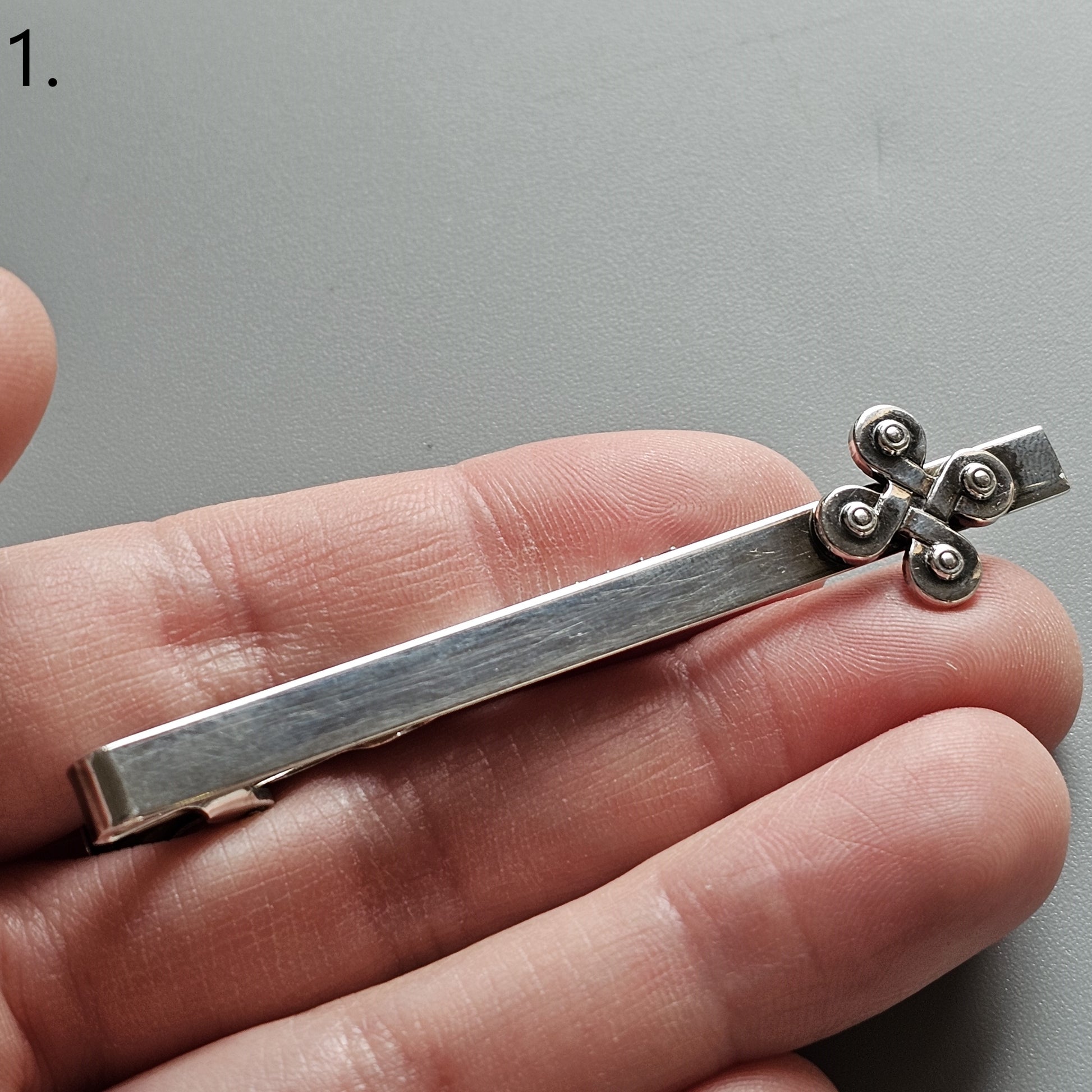 Silver tie clip with a decorative cross-shaped end adorned with small circular details.