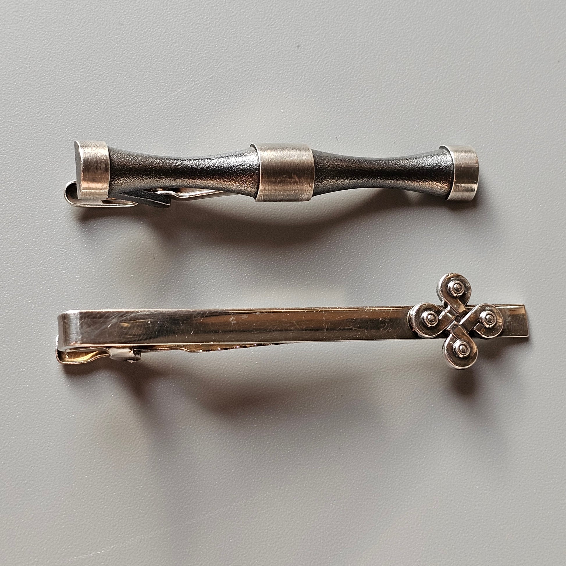 Two decorative metal tie clips or tie bars with different designs.