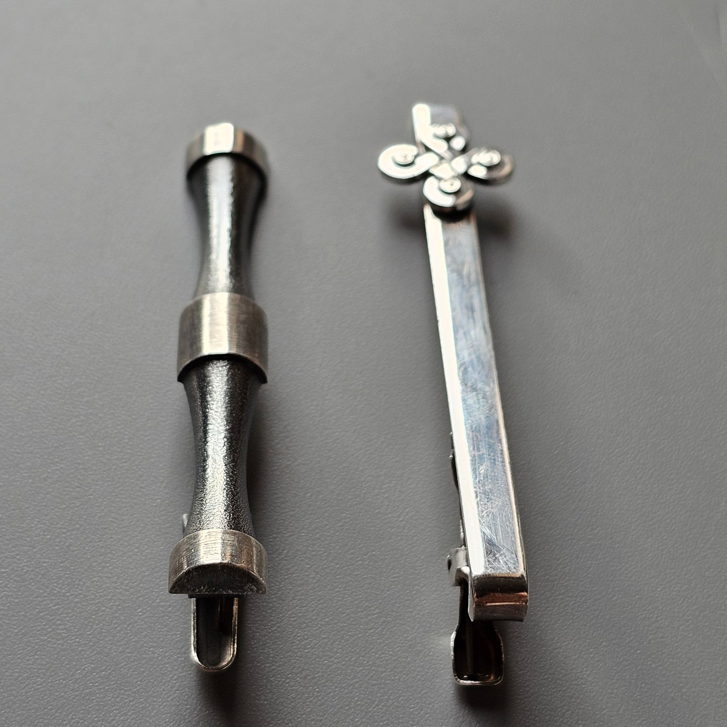 Two metal hair clips or barrettes, one with a plain cylindrical design and the other with a decorative floral shape at the end.