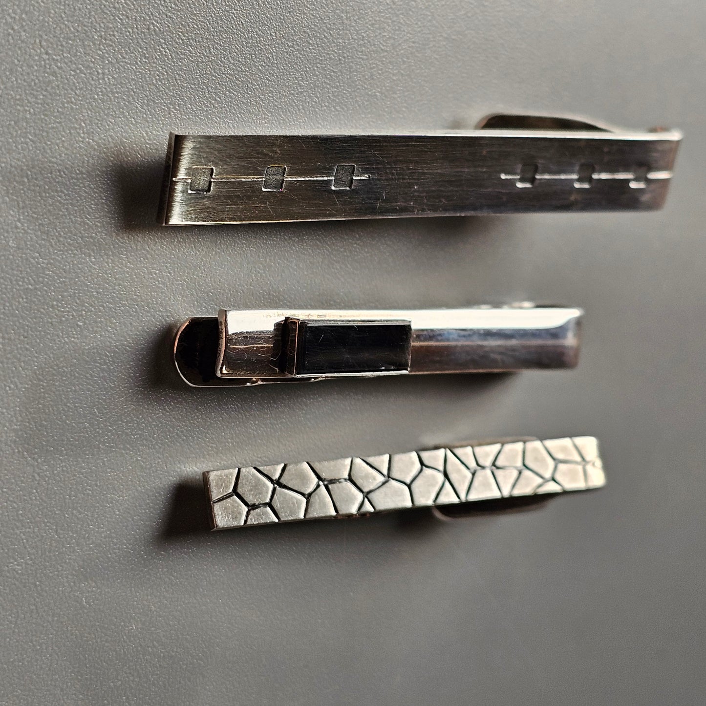 Three metallic tie clips with different designs and textures.