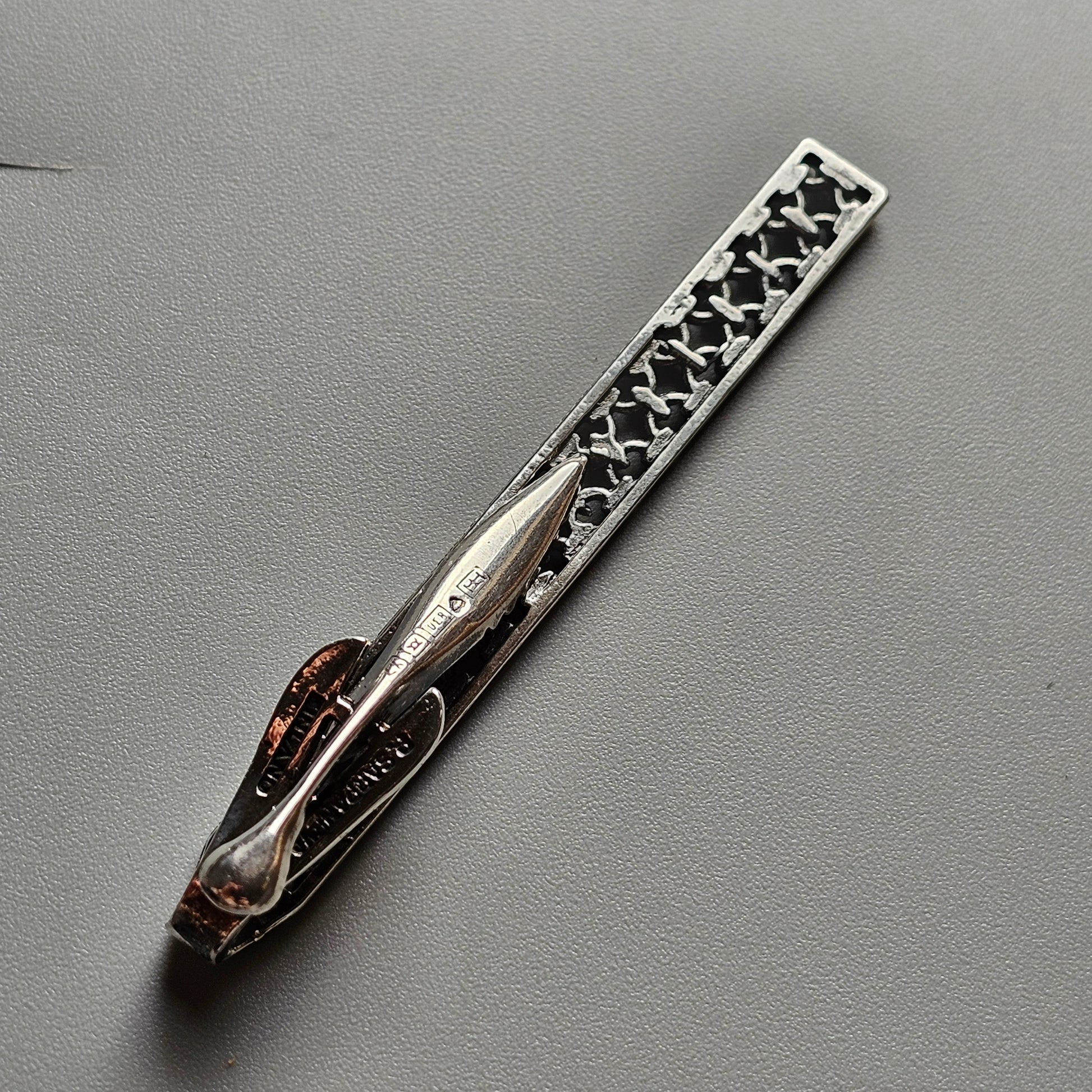 Ornate silver tie clip with intricate filigree design and a red gemstone accent.