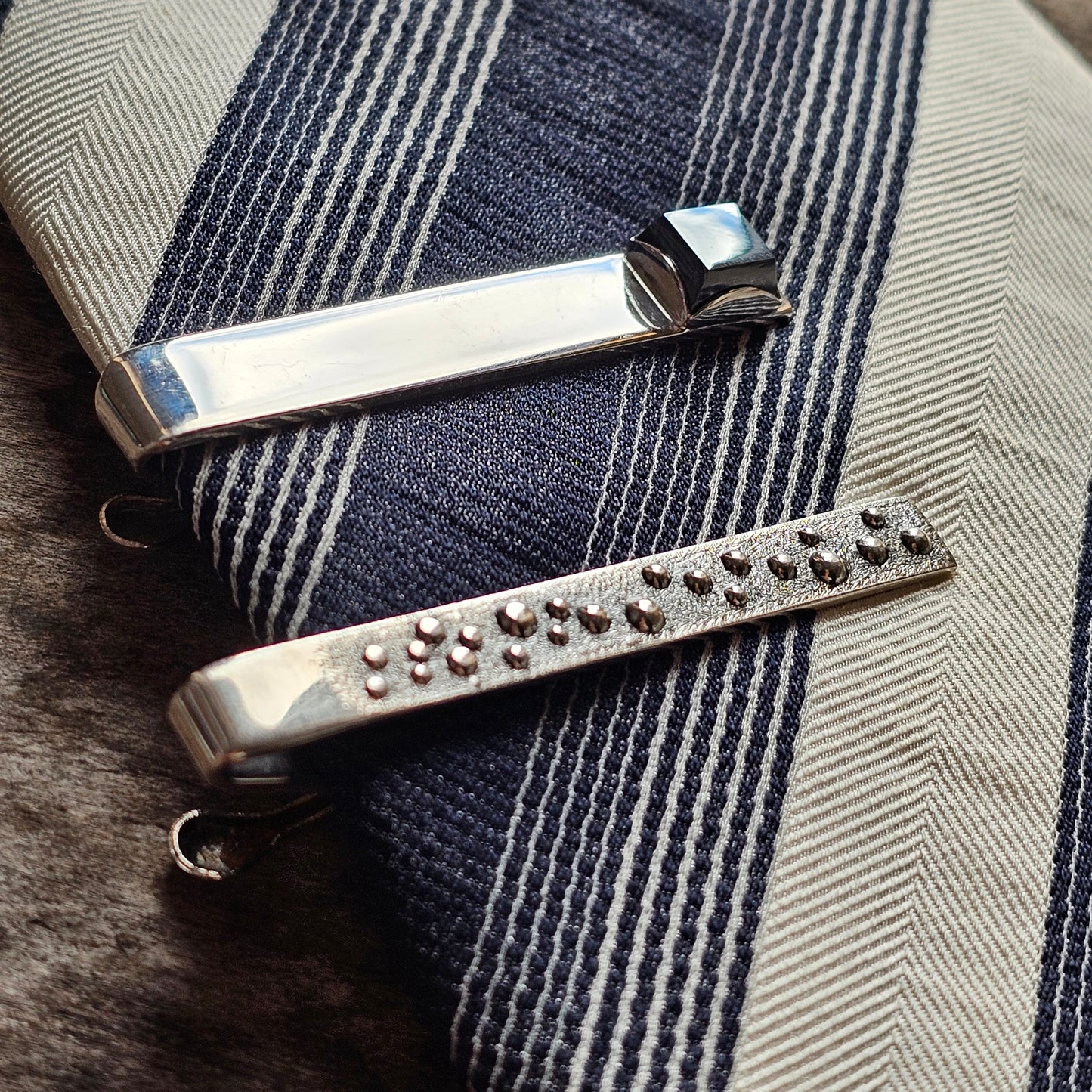 Two metal tie clips, one plain and one studded with small dots.