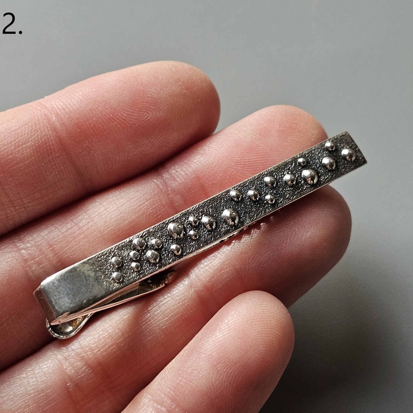 Metallic tie clip with a textured, studded surface design.